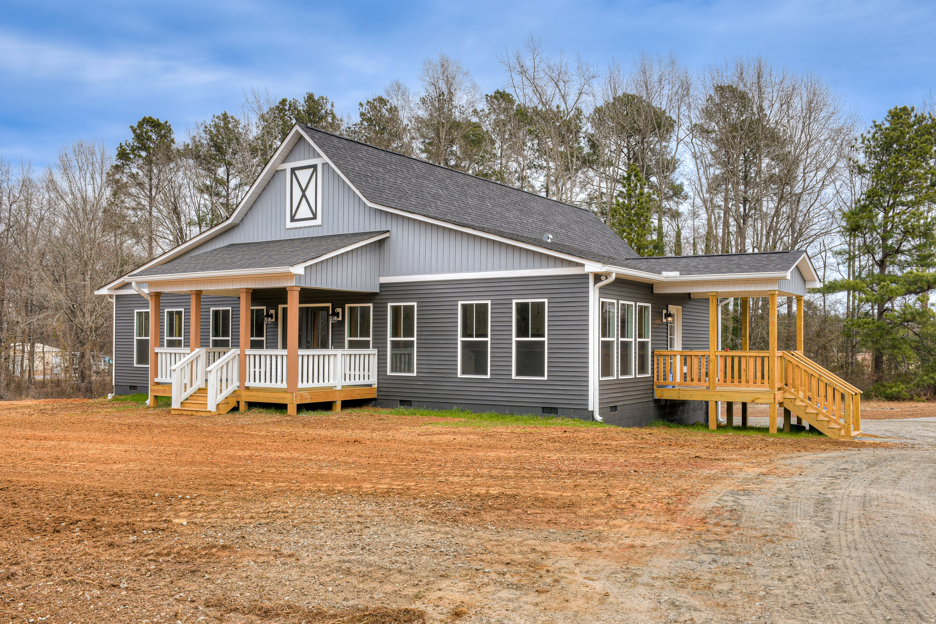 Two-story home with white siding, covered front porch, wooden stairs, concrete driveway, and dirt yard, surrounded by trees under a cloudy sky