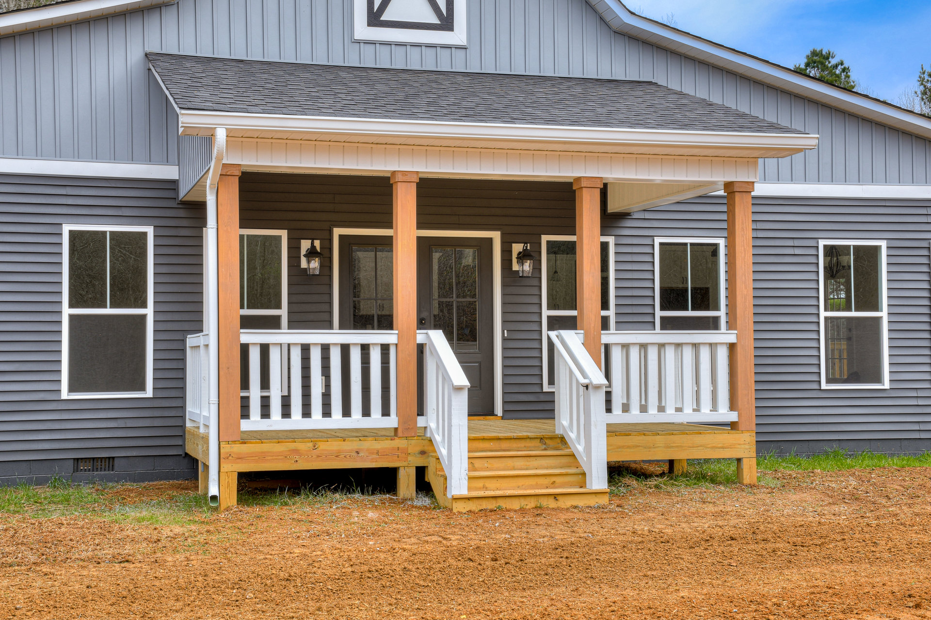 Two-story home with gray siding, covered front porch, white railings, wooden stairs, multiple windows, glass front door, and landscaped entry.