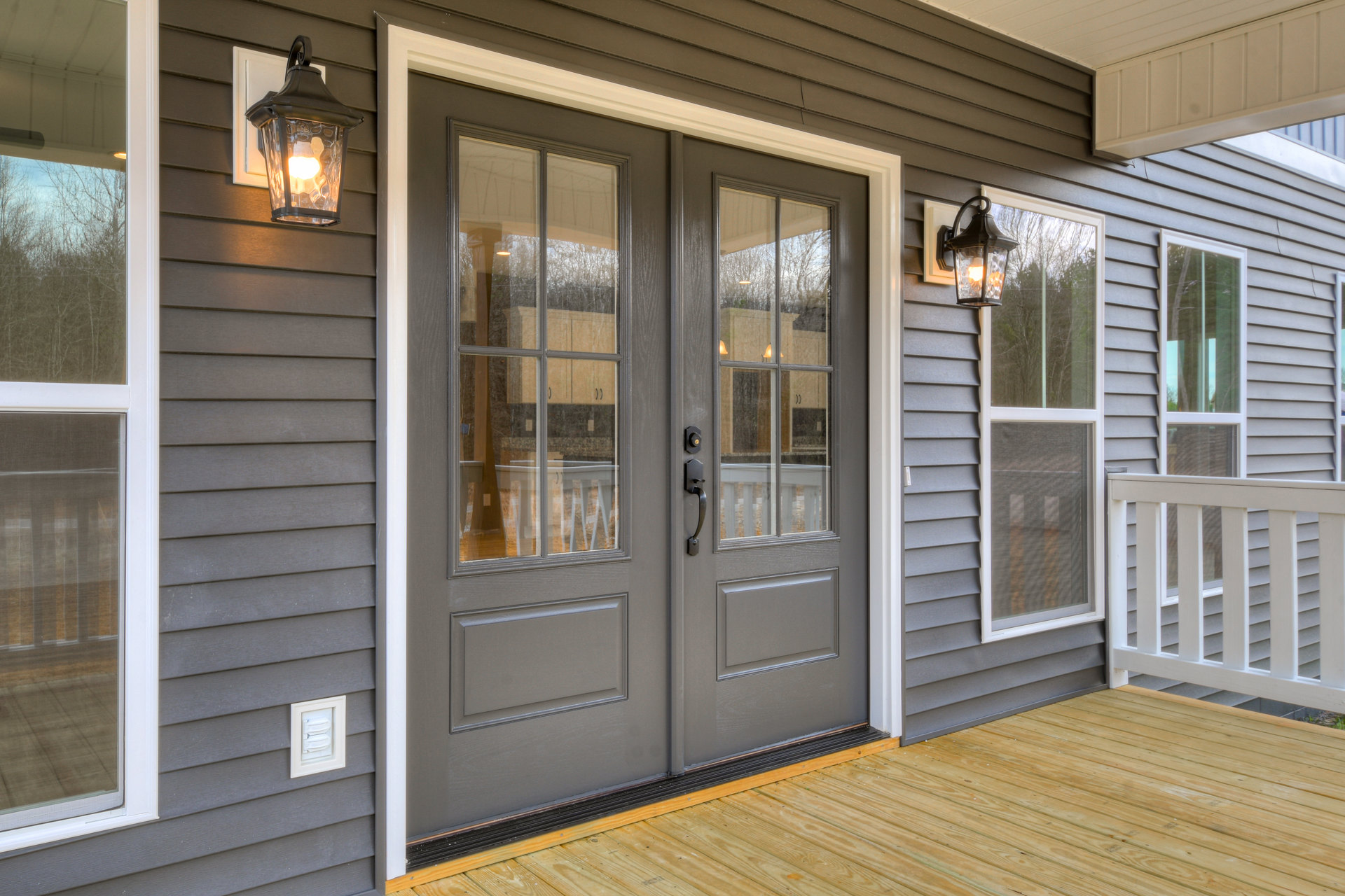 Double glass-paneled doors with white trim open onto a wooden deck, flanked by a white porch railing and illuminated by a modern light fixture.