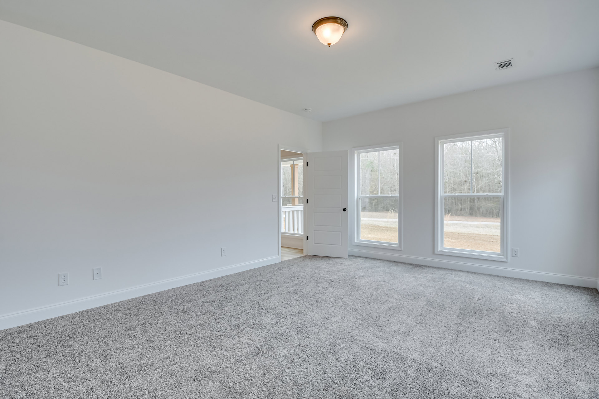Bright room with white walls, plush carpet flooring, black-knobbed white door, large windows revealing green trees, ceiling-mounted light fixture, and decorative molding.