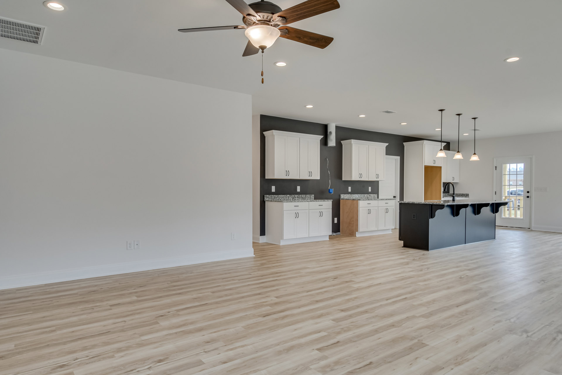 Open-concept room with hardwood flooring, ceiling fan with light fixture, kitchen area featuring white cabinets and black and grey countertops, door with window showing parked car