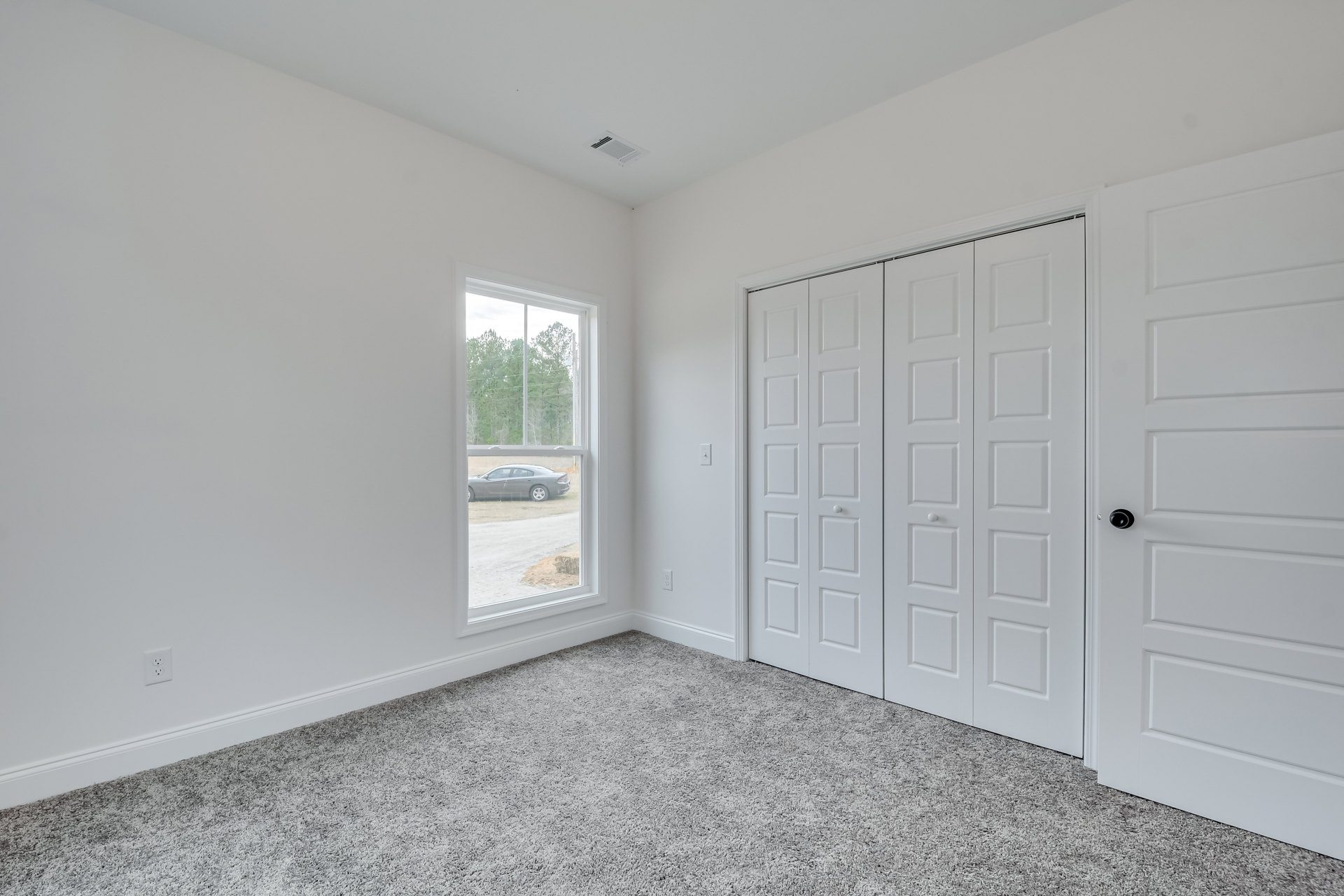 White-walled room with a carpeted floor, single door, and window overlooking trees and a parked car outside.