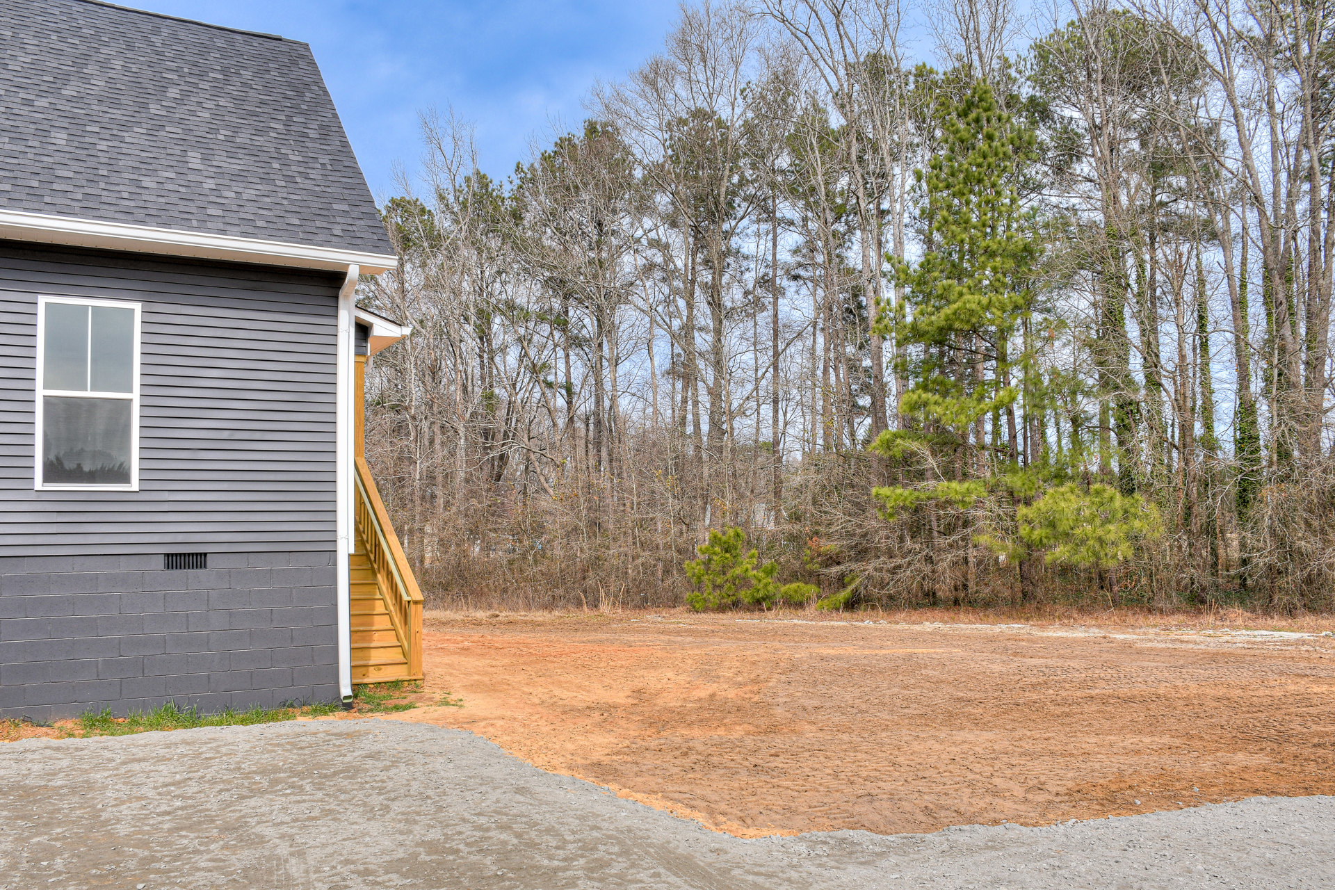 Two-story custom home with white-framed windows, wooden staircase and railing, paved driveway, and mature green trees in the background