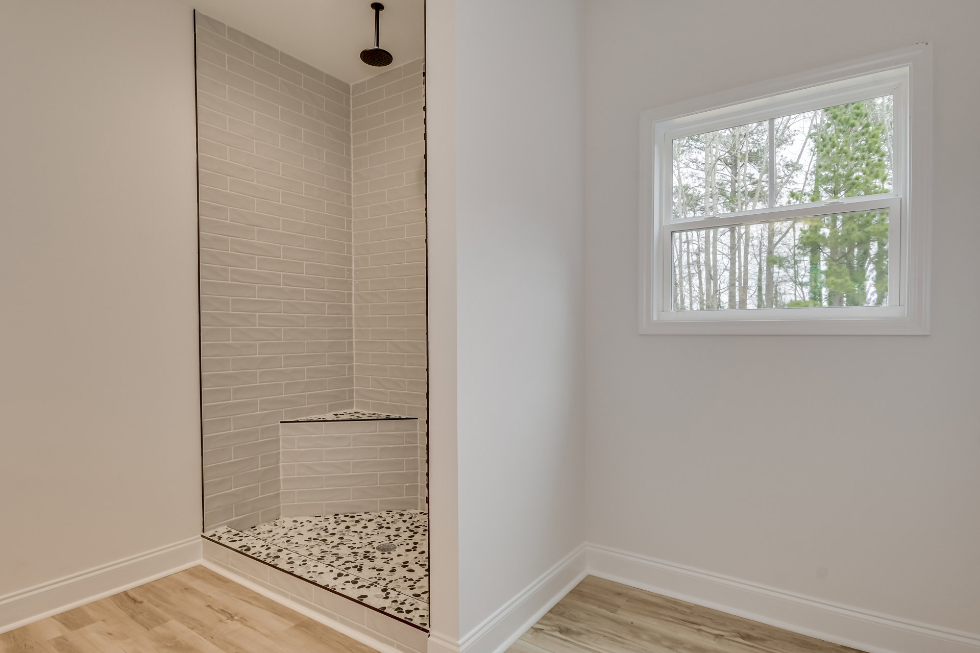 Walk-in shower with chrome showerhead, white tile walls, large window overlooking trees, wood flooring, and white baseboard trim