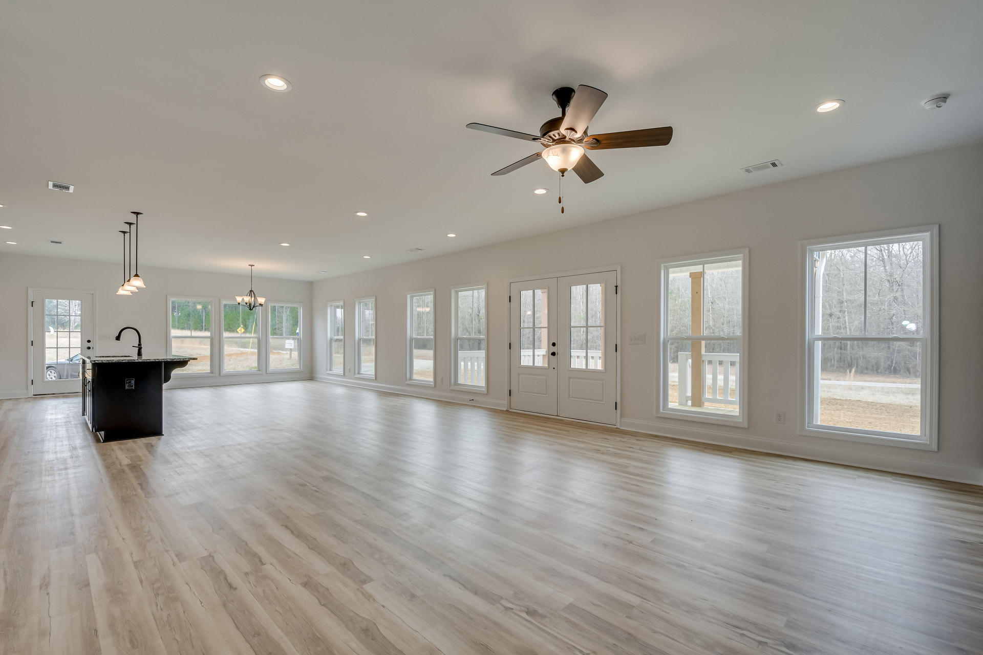 Spacious room with white walls, laminate wood flooring, ceiling fan with light fixture, double glass-paneled doors, large window showing trees outside, and black kitchen island