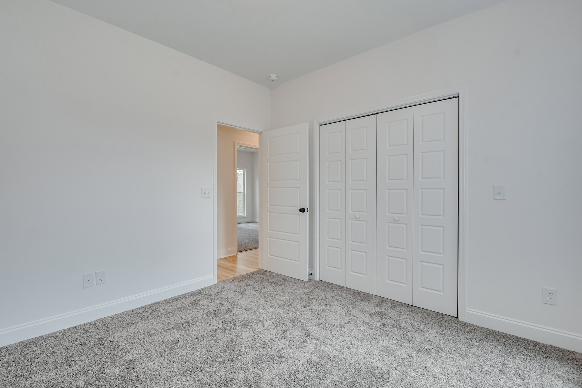 White-walled room with carpet flooring, black-knobbed white door, sliding closet doors, and window letting in natural light.