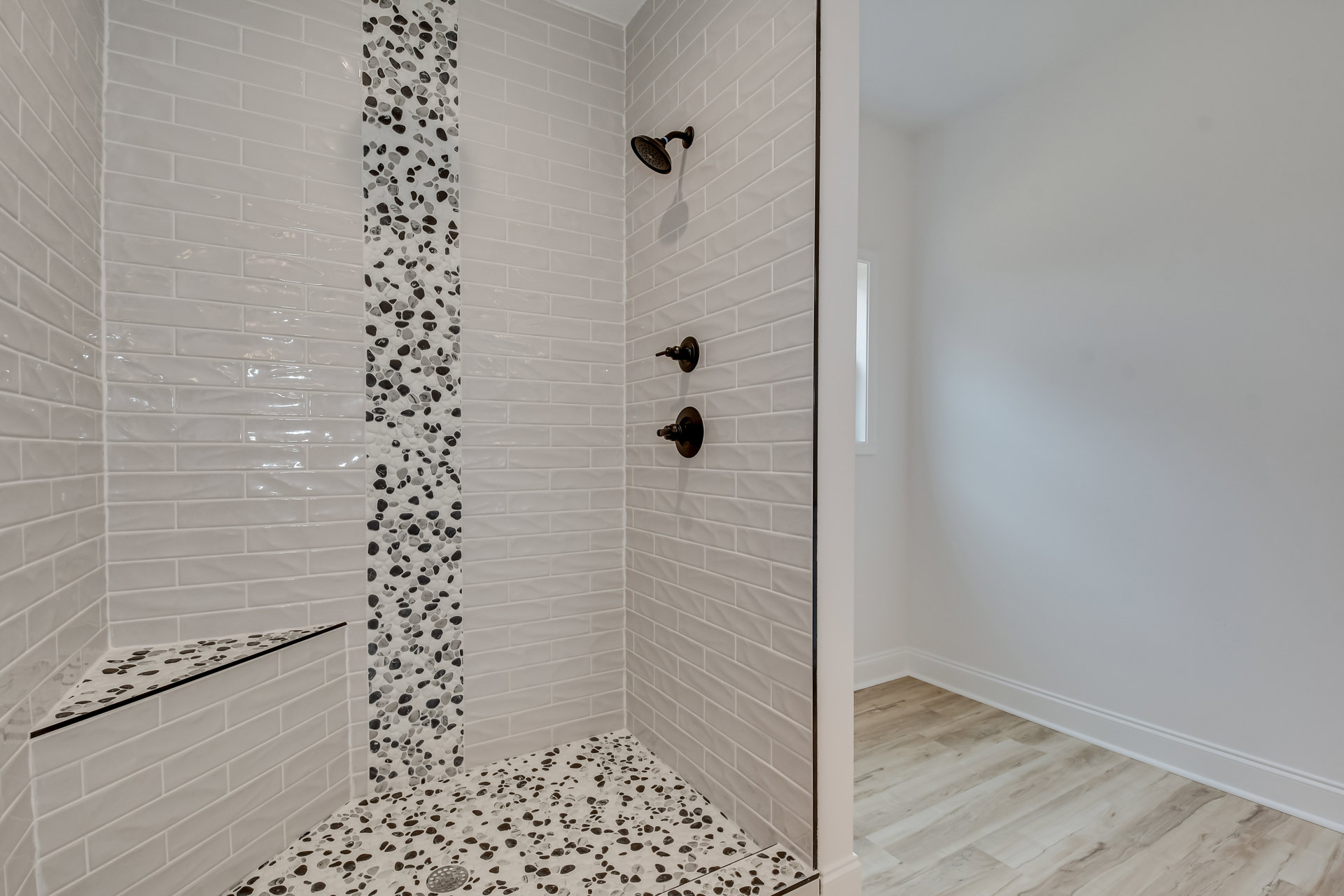 Walk-in shower featuring white tile walls, black and white pebble tile floor, central shower drain, black knob, and chrome shower head with blue accent band