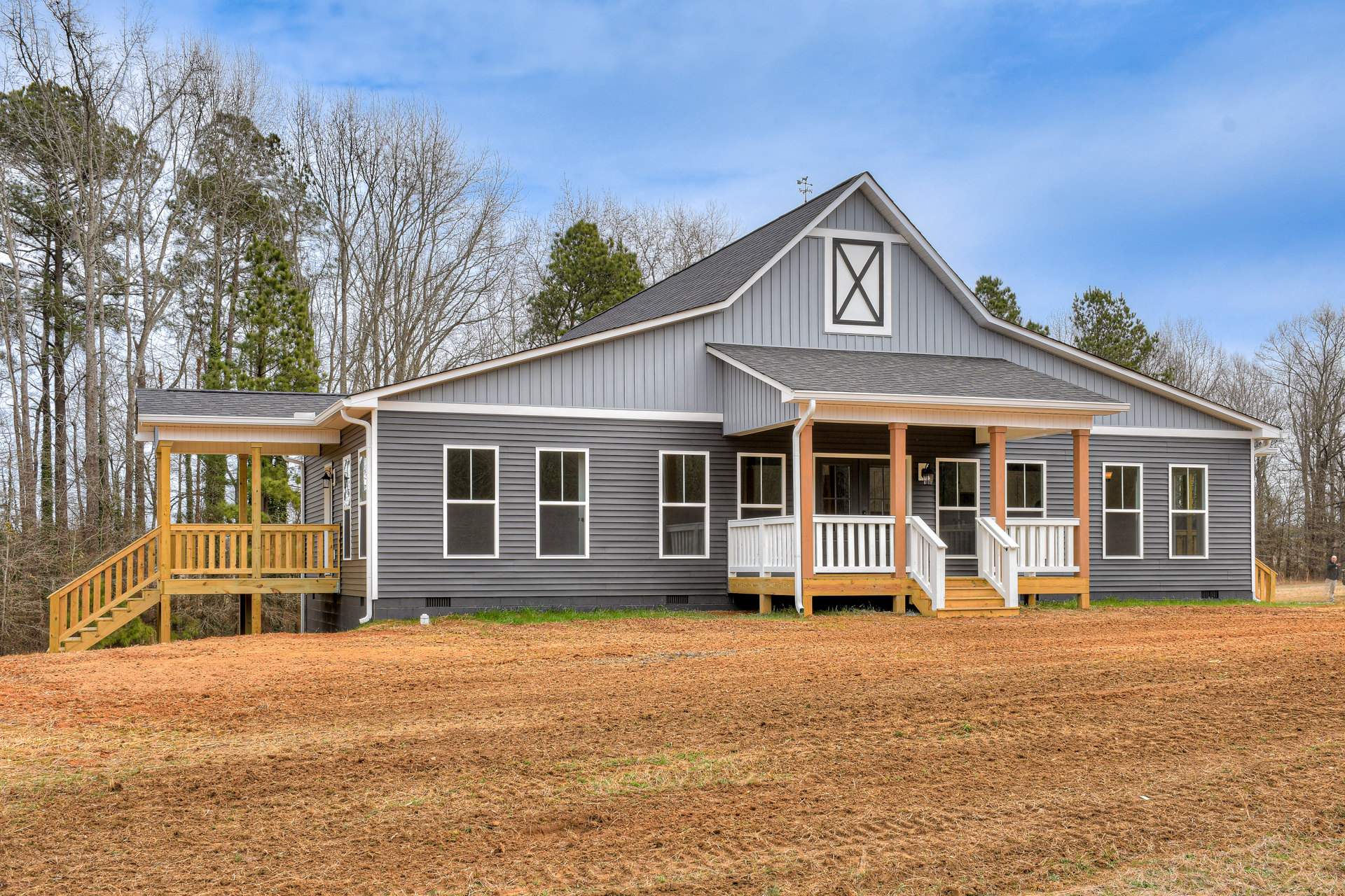 Grey cottage-style house with white-trimmed windows, wooden porch and stairs, expansive grassy yard bordered by trees under partly cloudy sky