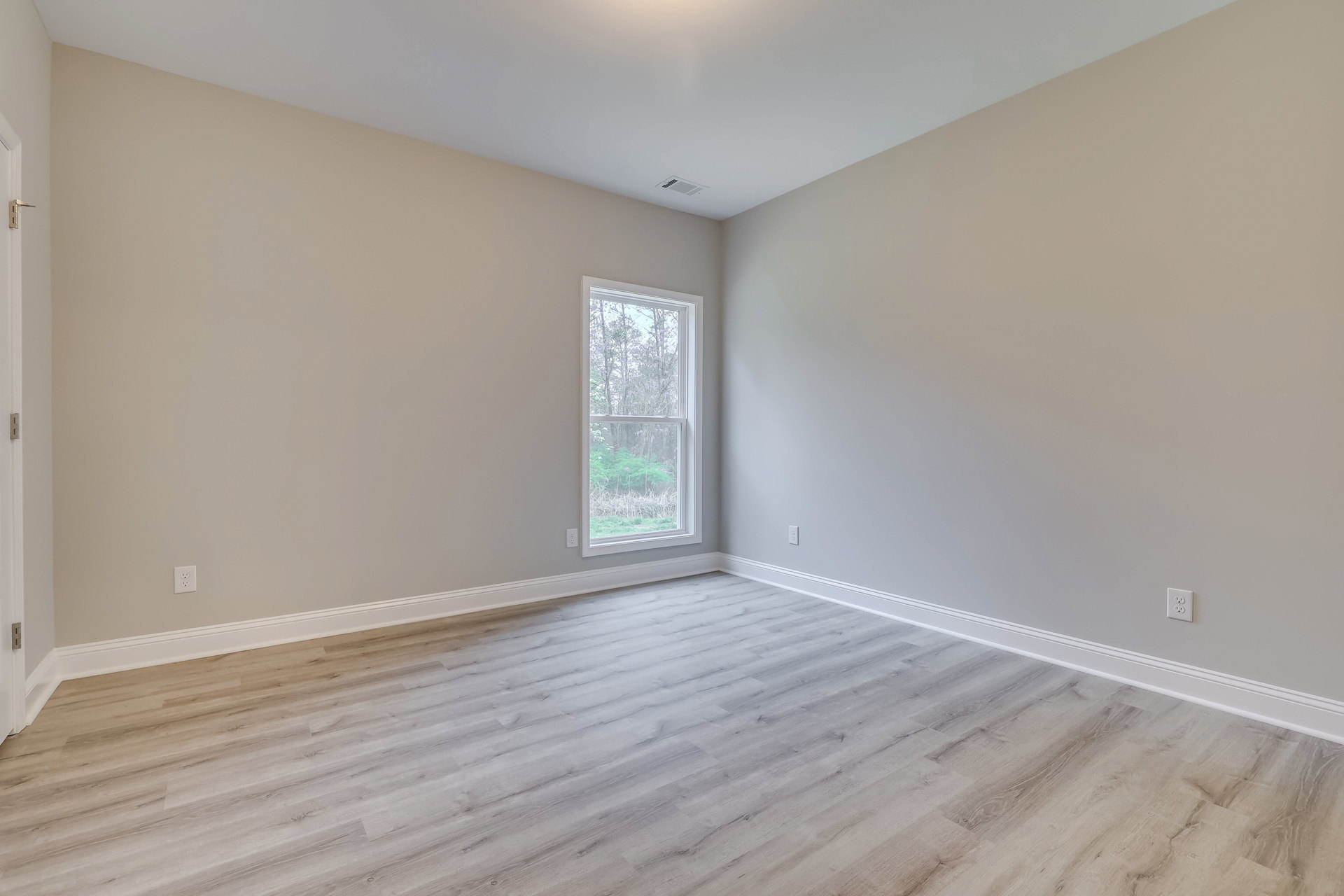 Sunlit room featuring a large window overlooking leafy trees, smooth hardwood flooring, white walls, and decorative molding.