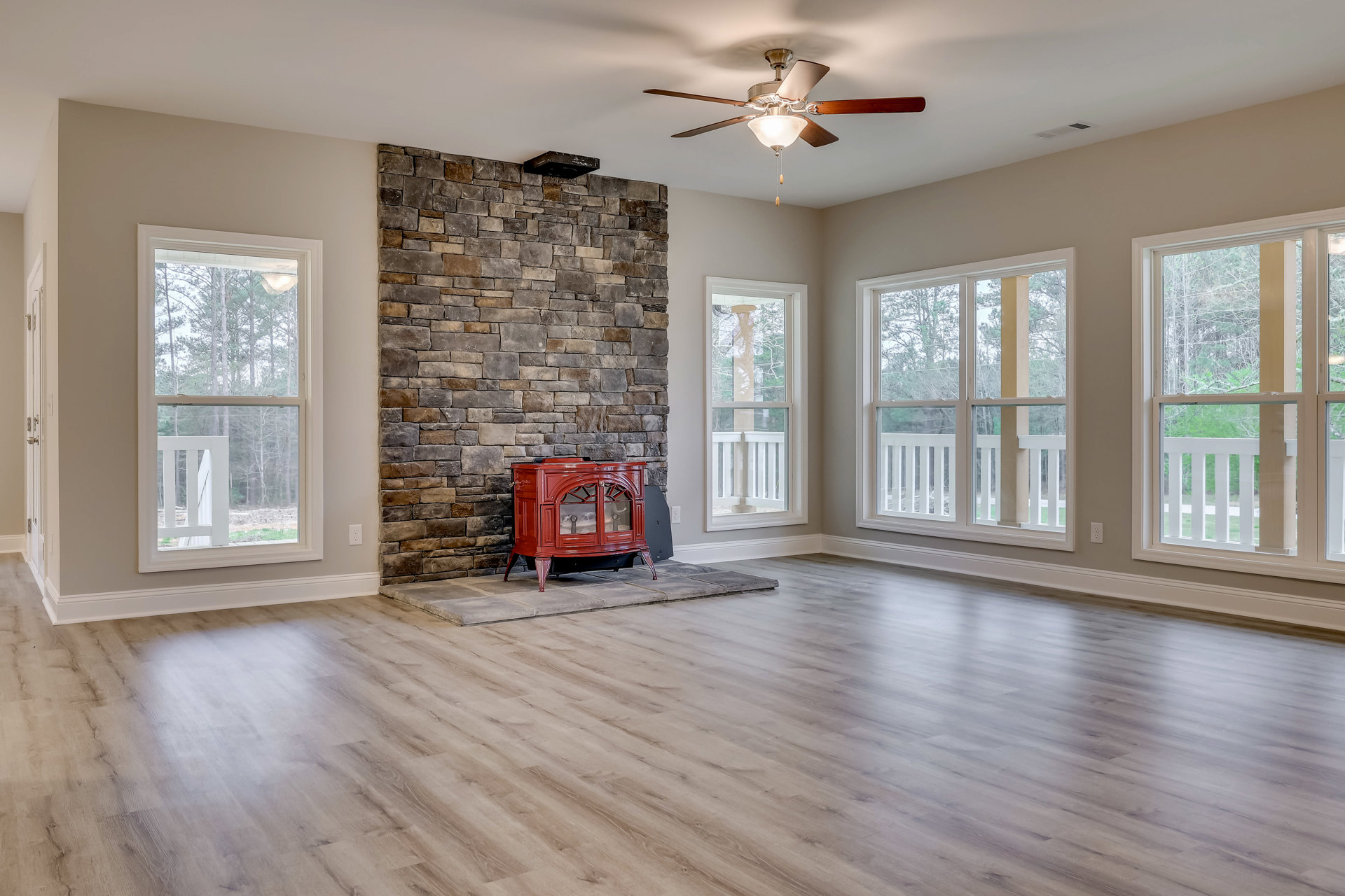 Living room with red wood stove fireplace, glass doors, wood flooring, ceiling fan with light fixture, large window showing trees and white railing outside