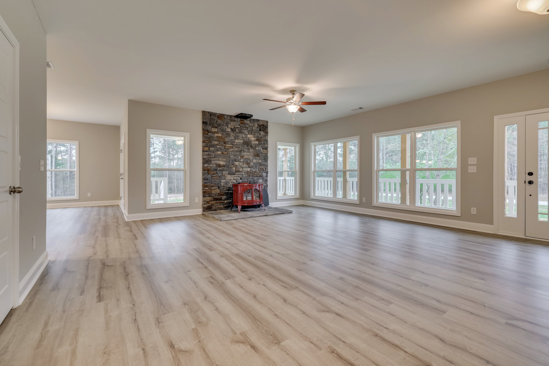 Living room with wood flooring, red wood stove fireplace with glass doors, ceiling fan with light fixture, large window showing trees outside