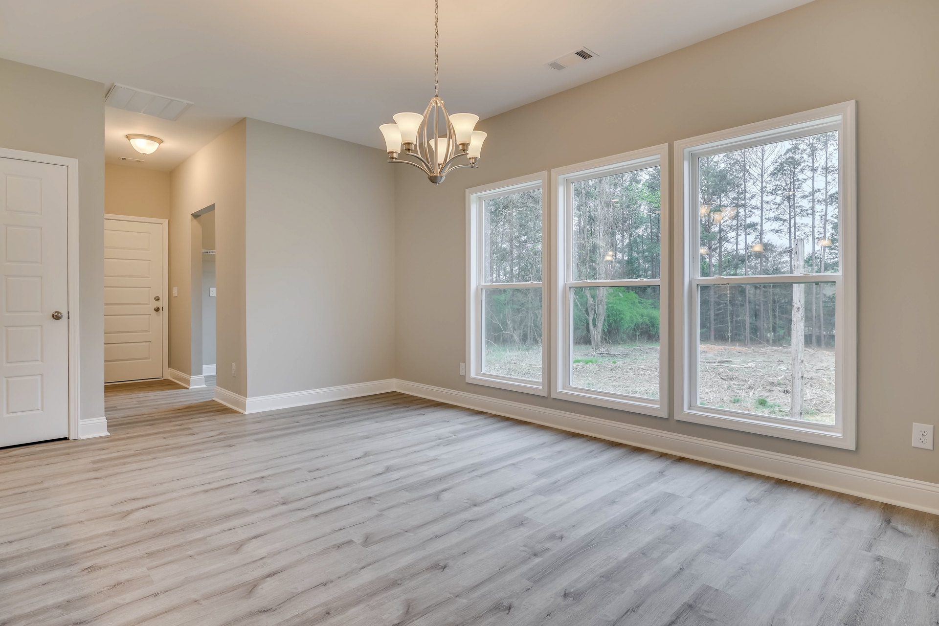 Spacious room with wood flooring, multiple large windows showing trees outside, white door with silver handle, ornate five-light chandelier hanging from ceiling, white walls with