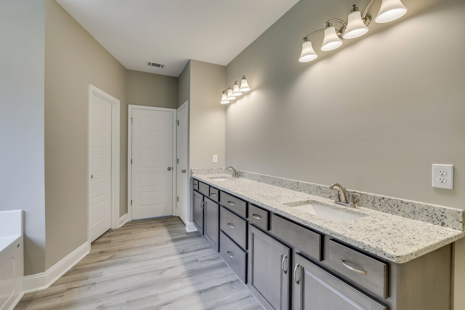 Bathroom with double sink vanity, wood flooring, white cabinetry, silver faucet, three-light fixture above mirror, white door with silver knob, and white electrical outlet on wall.