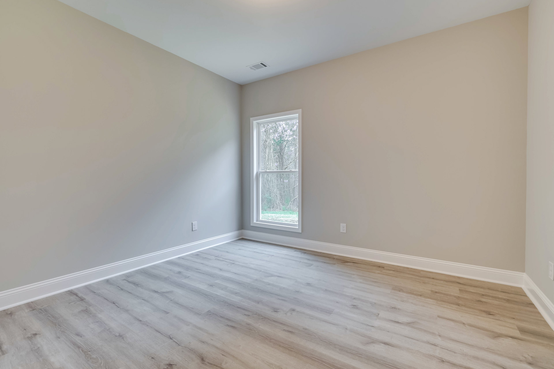 Sunlit room featuring wide-plank hardwood flooring, white plaster walls, ceiling vent, large window overlooking leafy trees, and simple base molding.