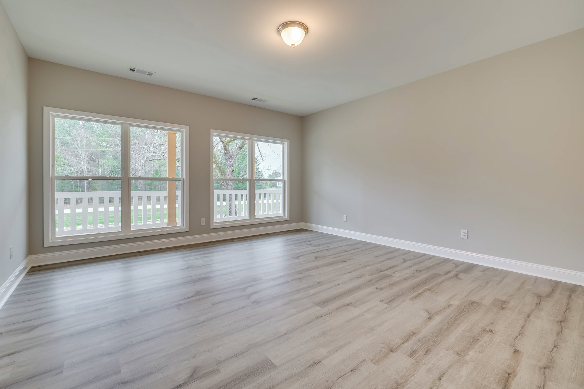 Sunlit room with wood flooring, large windows overlooking trees, ceiling light fixture, and adjacent porch visible through glass doors