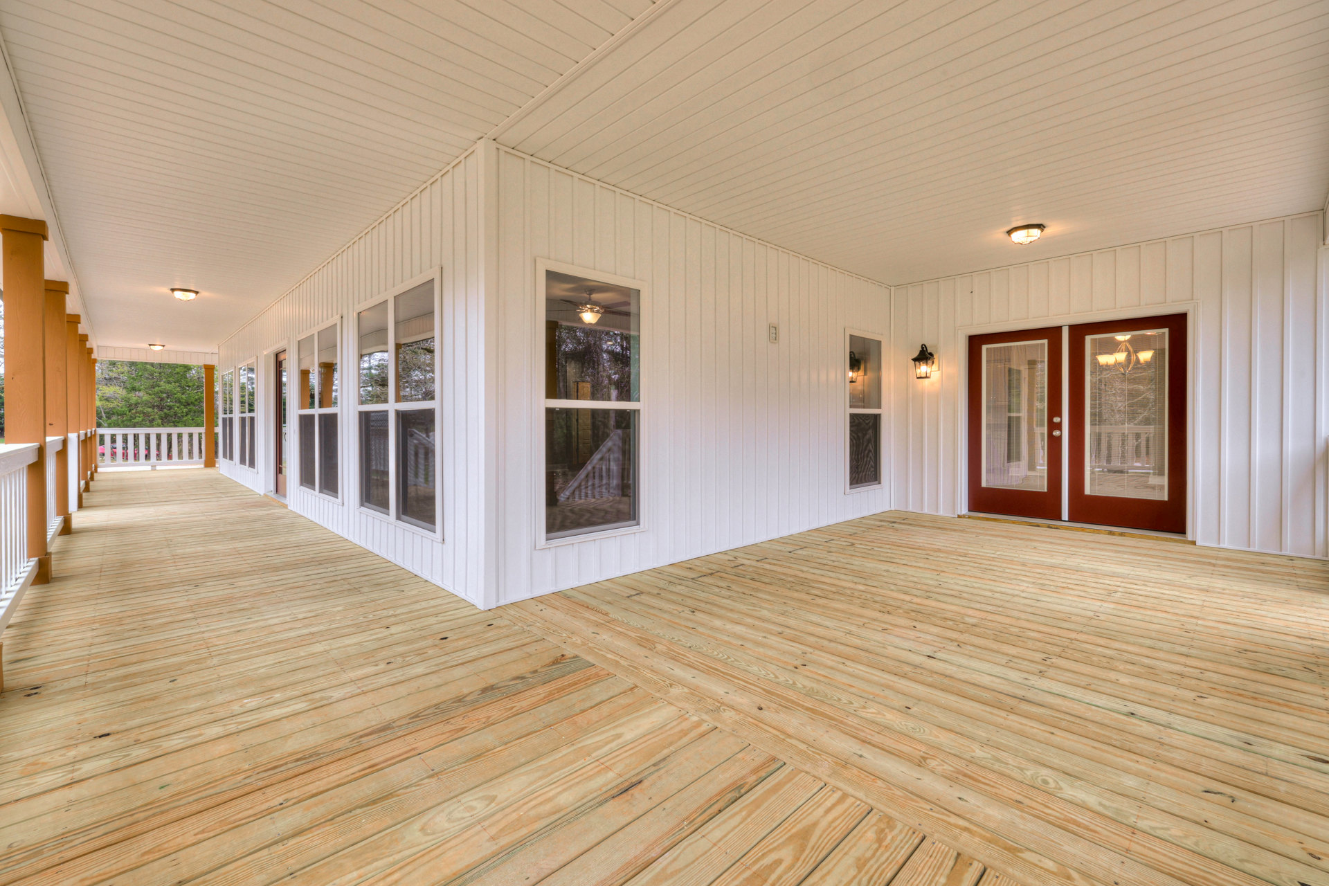 Spacious white home interior featuring wood flooring, double doors with blinds, large windows overlooking trees, staircase, wooden deck, and modern light fixture.