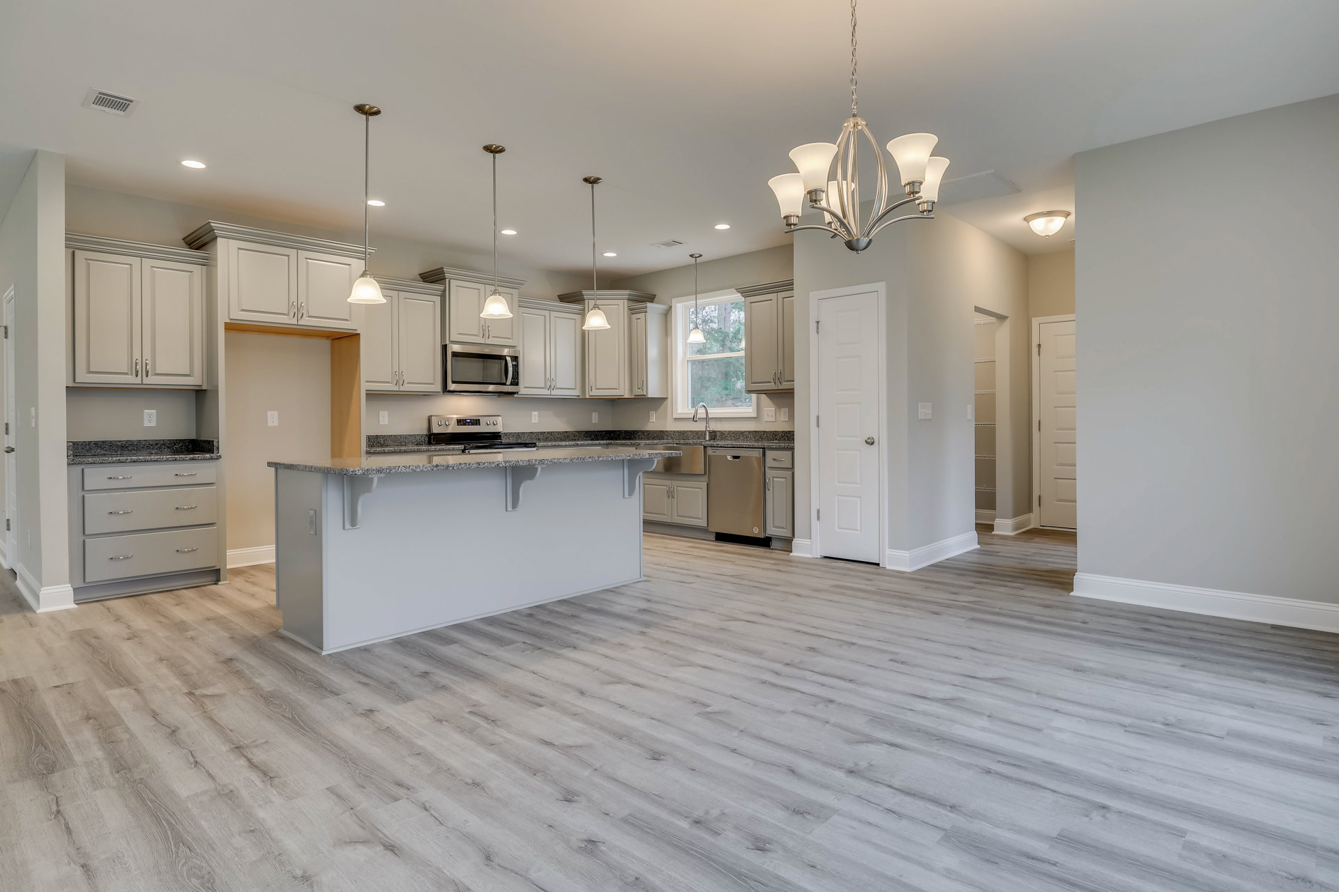 Open kitchen and dining area featuring wood flooring, white countertops with drawers, black-door microwave, modern chandelier, white door with silver handle, and light-colored