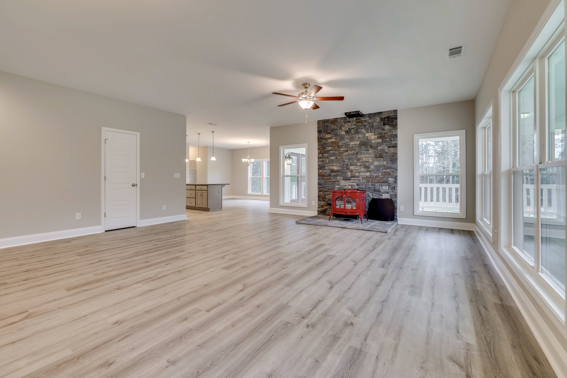 Spacious living room featuring wood laminate flooring, stone fireplace with glass-fronted red stove, ceiling fan with light, white door with silver handle, large window overlooking