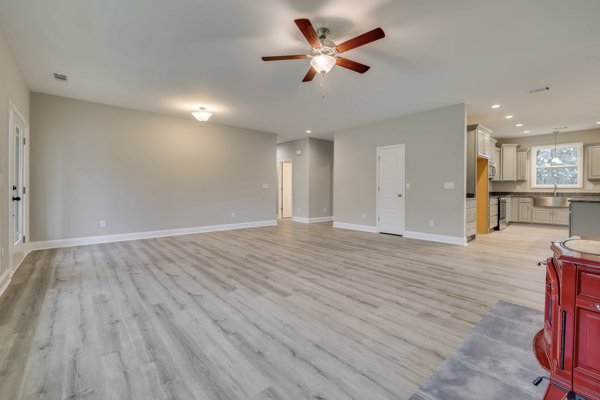 Spacious room featuring hardwood flooring, white walls, ceiling fan with light fixture, white door with silver handle, and red metal object near the wall