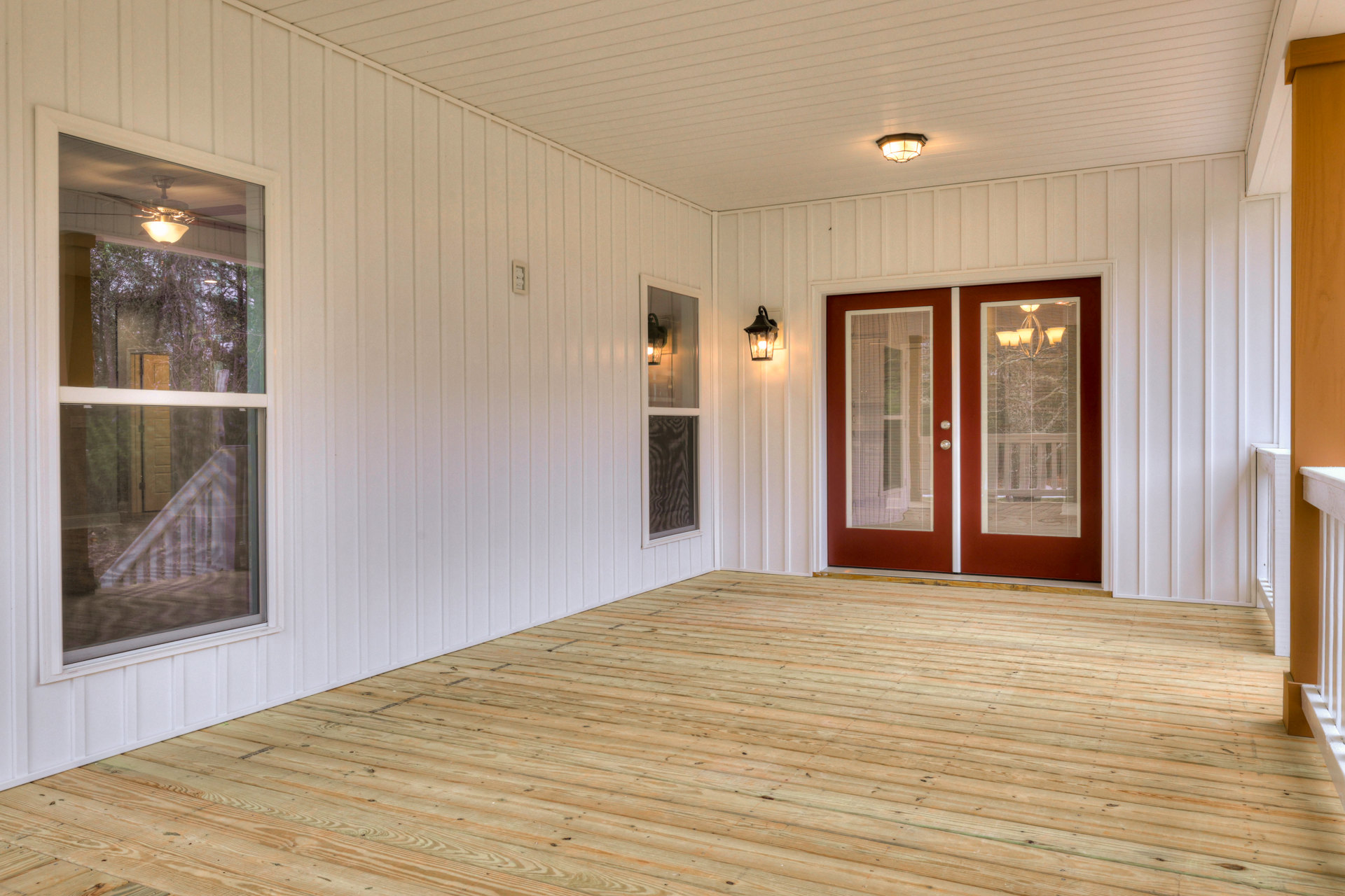 White walls and wood flooring in a bright interior, double red doors with blinds, ceiling and wall light fixtures, white railing visible outside window.