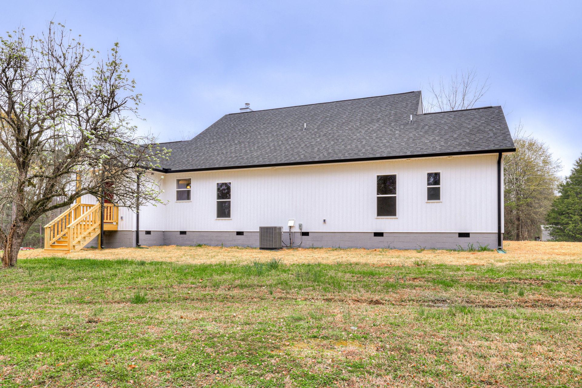 White house with black roof, large front window, green lawn, leafy tree in foreground, partly cloudy sky
