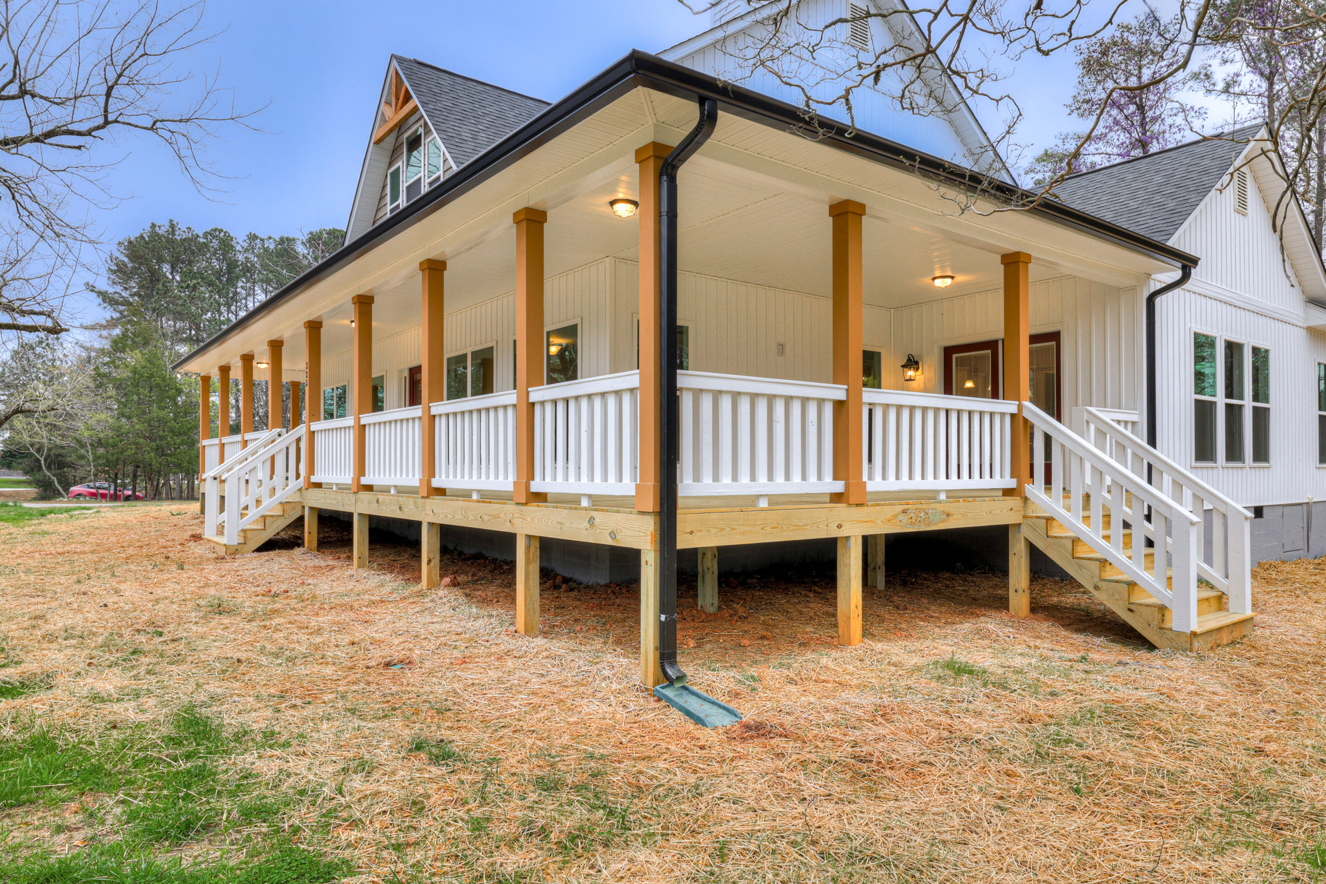 Two-story house with light siding, large wooden porch featuring white railings, visible gutters along the roofline, and mature trees in the background