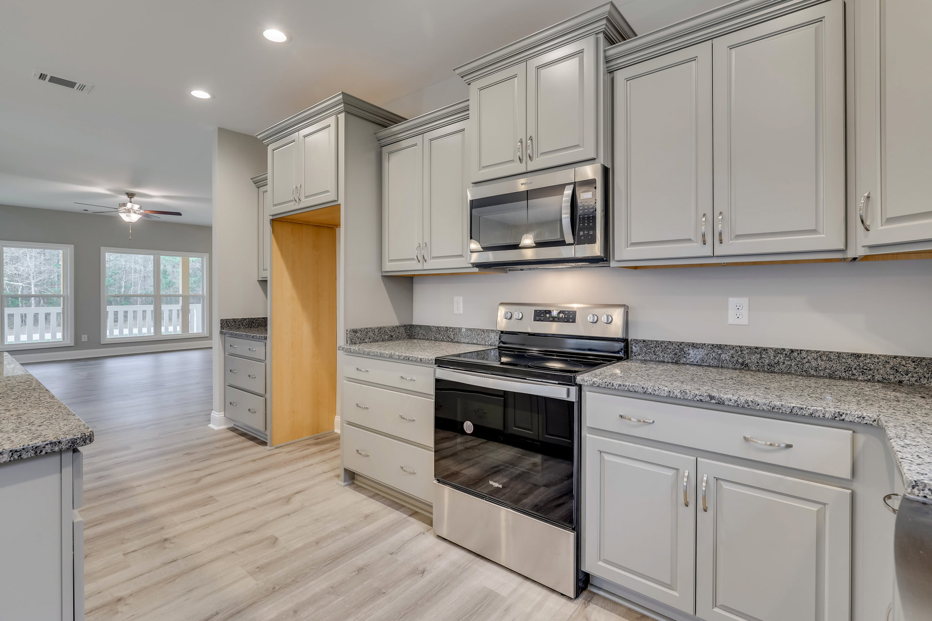 White kitchen with silver-handled cabinets, stainless steel stove and oven, built-in microwave, ceiling fan with light, window overlooking trees, light countertops.