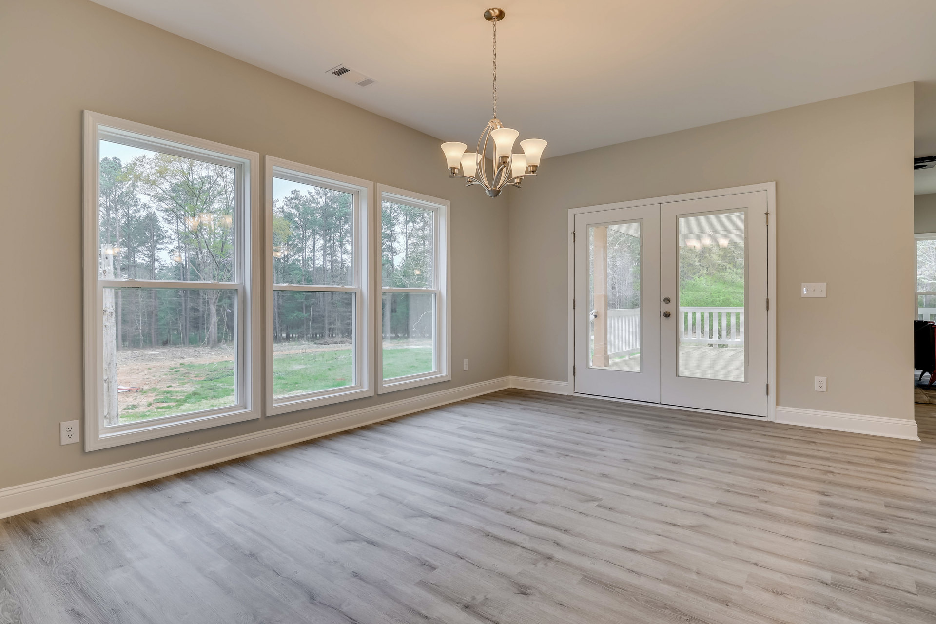 Spacious room with wood flooring, large windows overlooking trees, glass-paneled double doors, and a decorative chandelier hanging from a white ceiling with crown molding