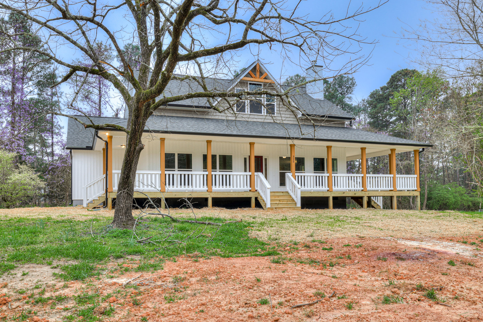 Spacious wooden porch with white railing, large tree with moss in the background, light-colored siding, multiple windows, manicured lawn