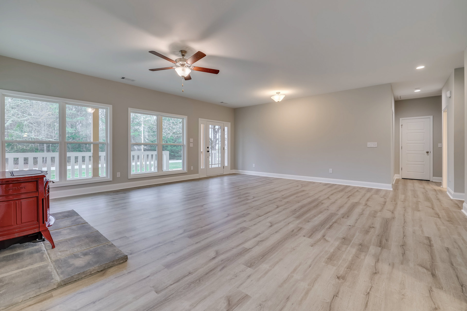 Open room with wood plank flooring, white walls, ceiling fan with light fixture, white door with silver handle, glass-paneled door revealing tree outside, red metal object with