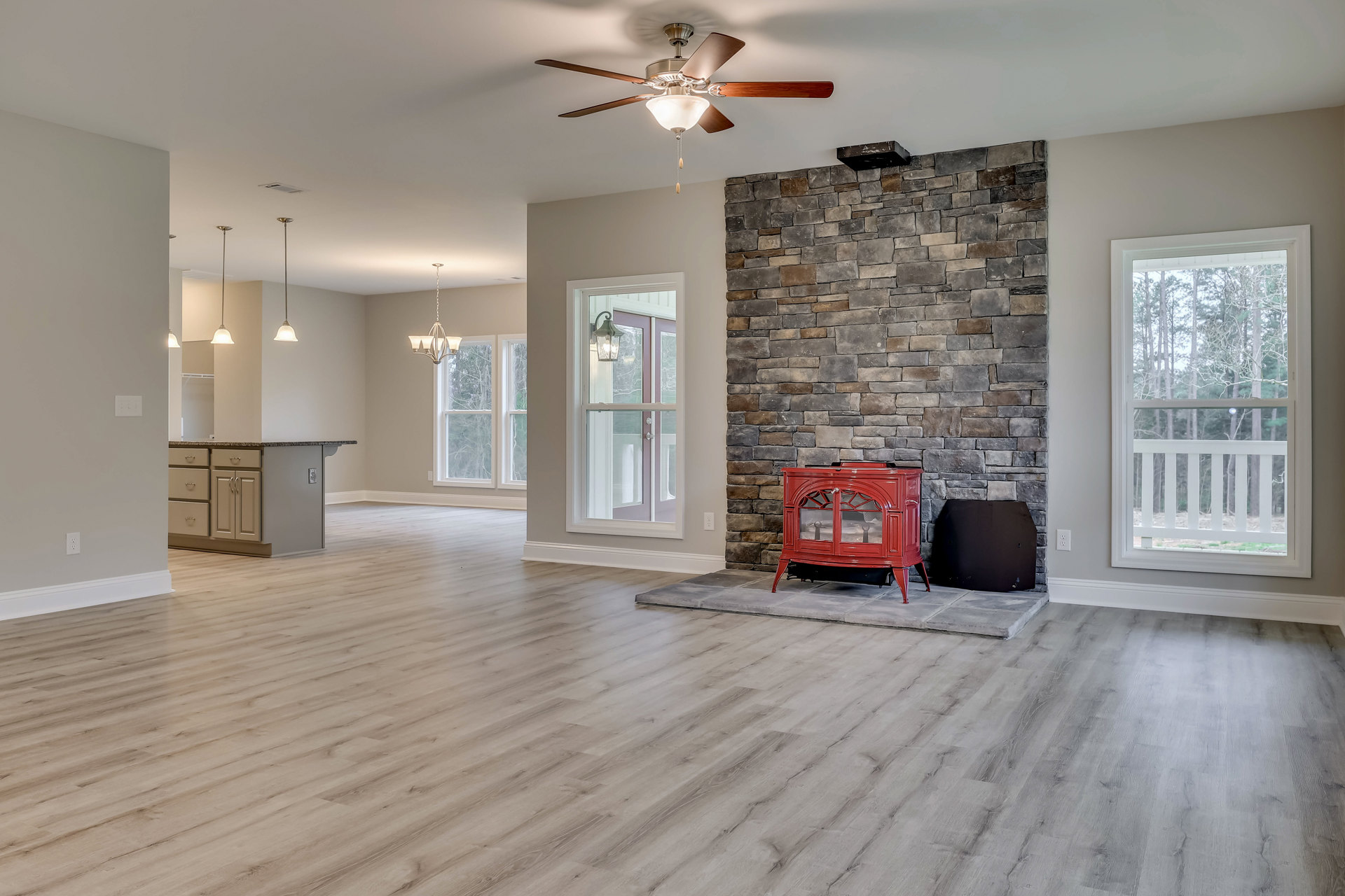 Living room with wood flooring, red stove fireplace with glass doors, large window showing trees outside, ceiling fan with light fixture, plaster walls