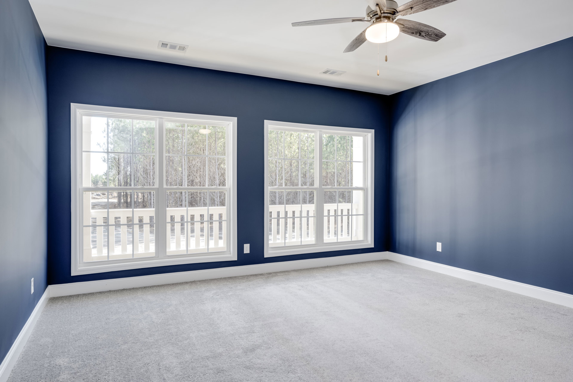 Blue accent wall with two white-trimmed windows, ceiling fan with light fixture, and white carpet flooring