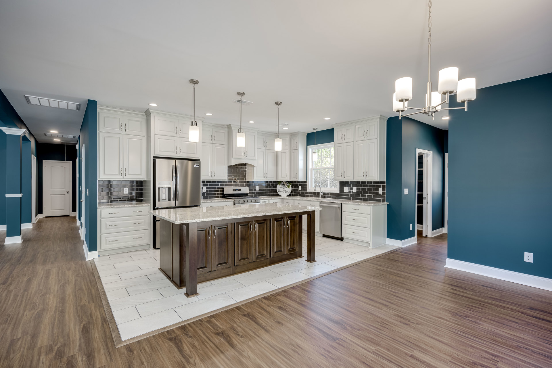 Kitchen with wood flooring, central island featuring marble countertop, silver refrigerator, white door with silver knob, cabinetry, and decorative wall sign with bow; close-up of