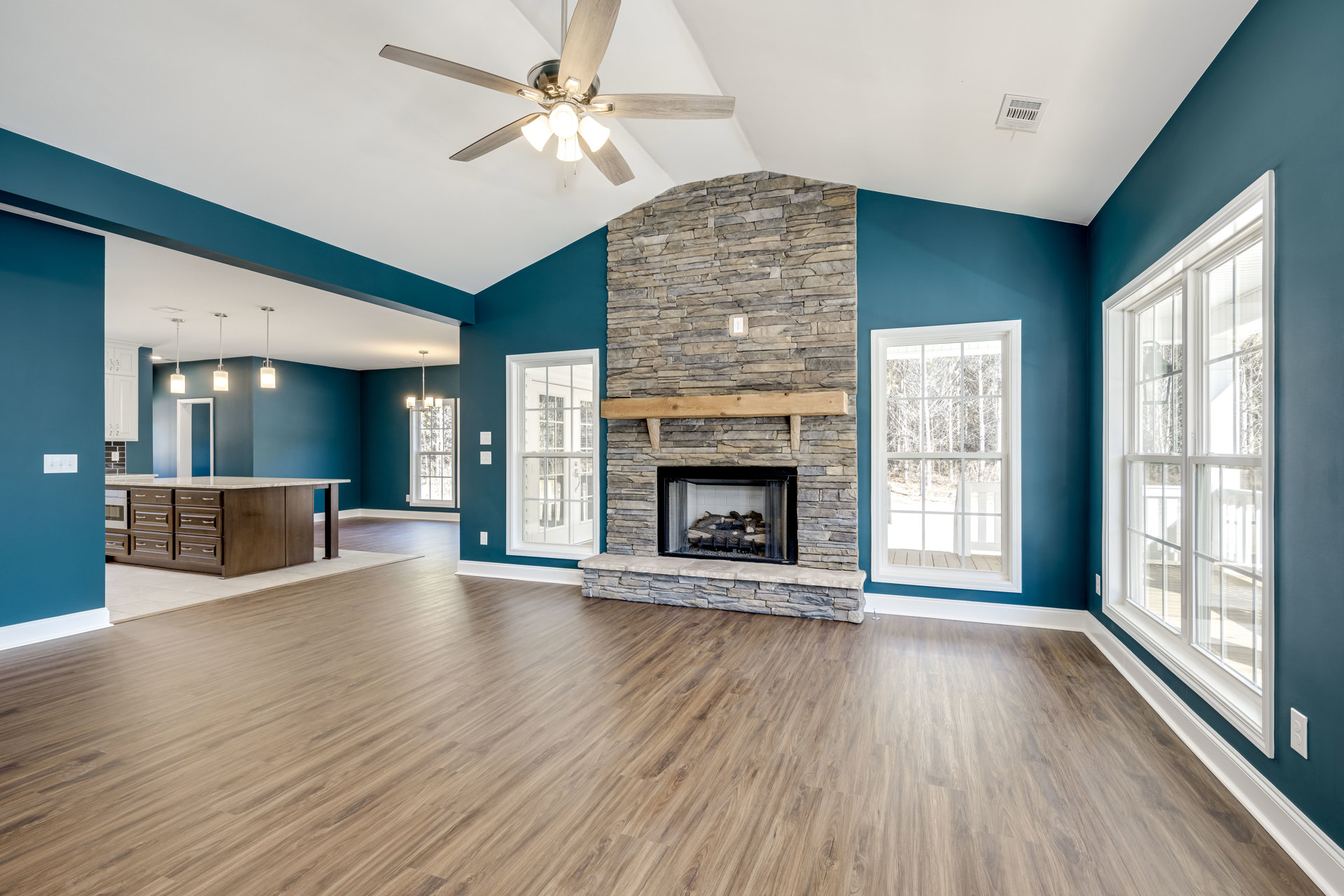 Living room with hardwood flooring, ceiling fan with light fixture, fireplace stacked with logs, large window showing trees outside
