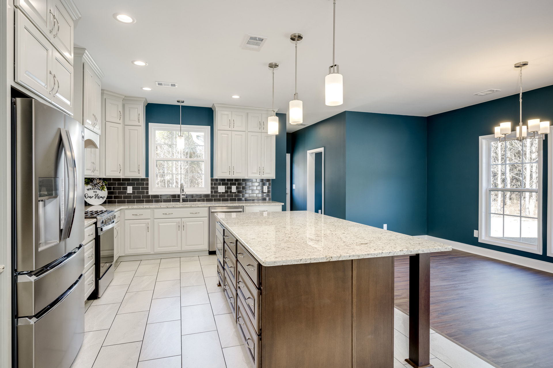 Spacious kitchen featuring a large marble-topped island, white cabinetry, stainless steel refrigerator, tile backsplash, and a window overlooking trees.
