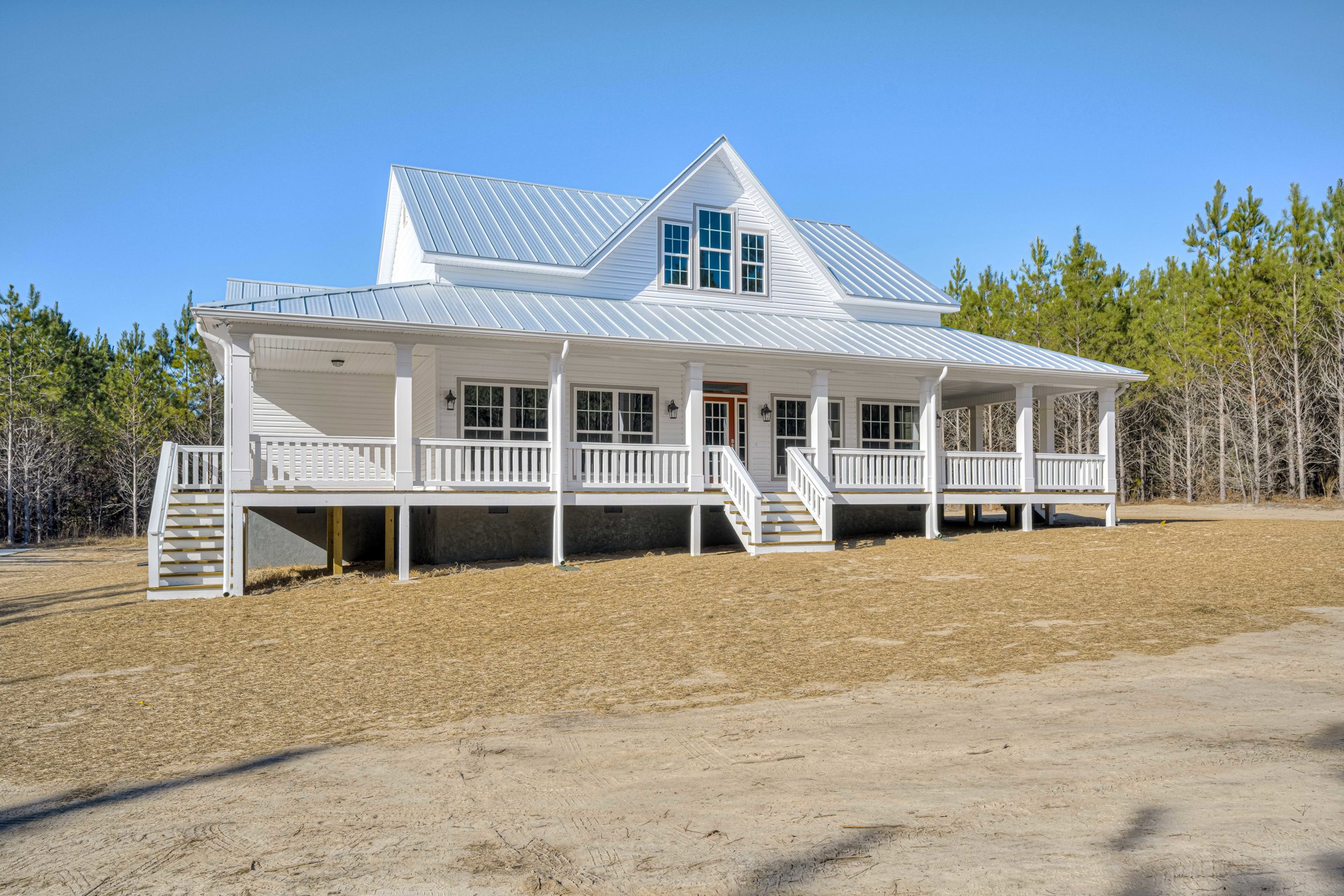 White siding house with covered porch, white railings, multiple windows, and gabled roof, surrounded by trees under blue sky