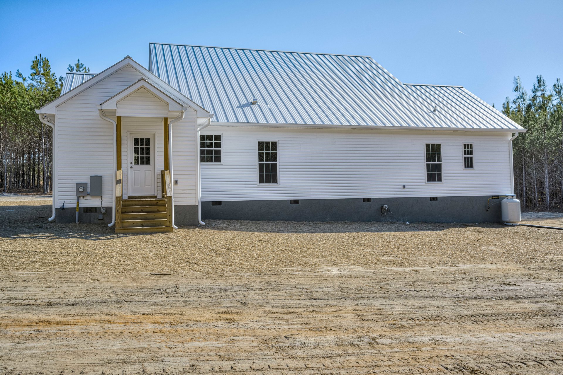 White house with white siding and white-framed windows, gable roof, dirt field in foreground, white pipe and capped cylinder near dirt road, sparse trees in background.