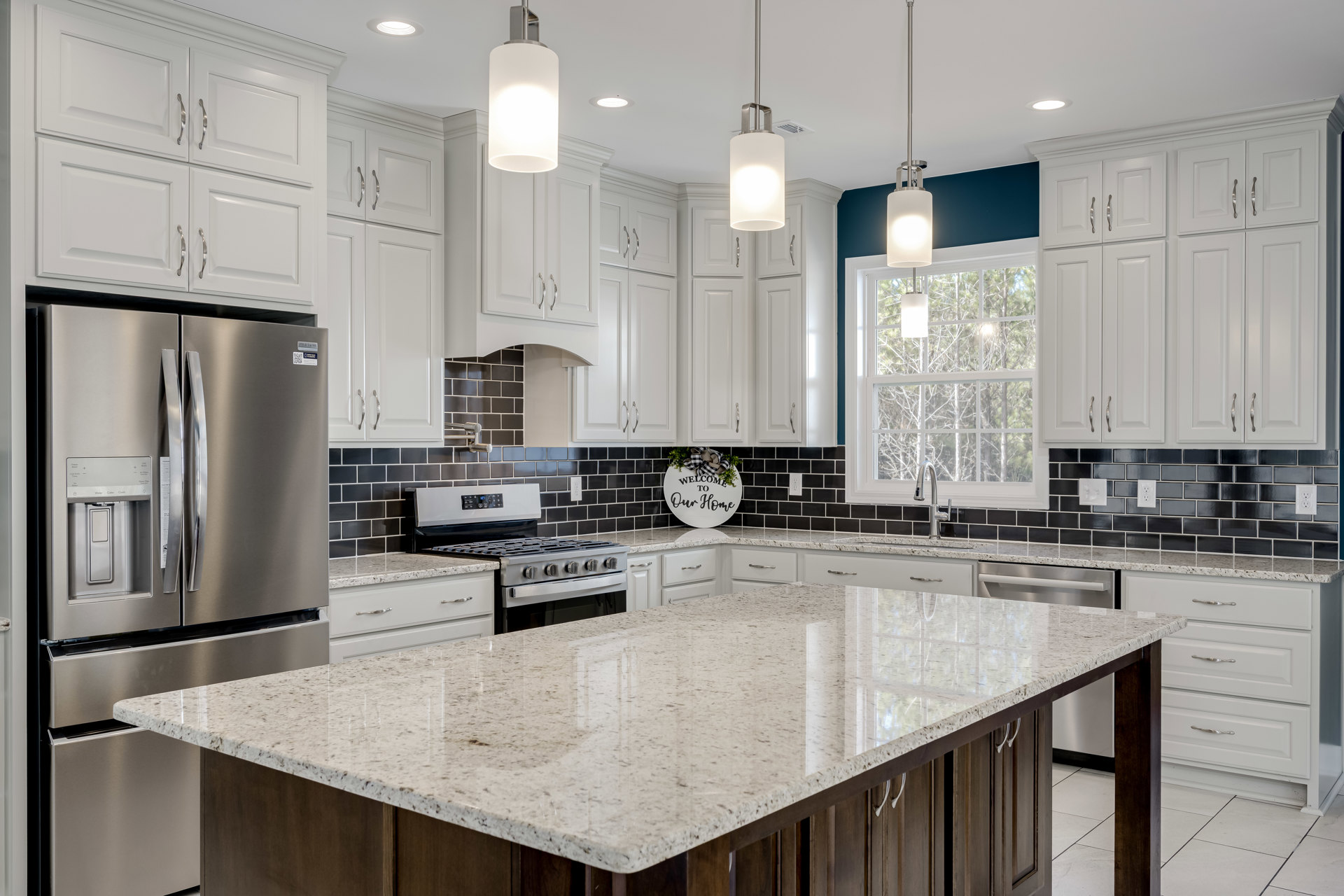 White kitchen with marble countertops, central marble island with built-in stove, stainless steel refrigerator, white cabinetry, wall-mounted sign with bow, sword holder displayed