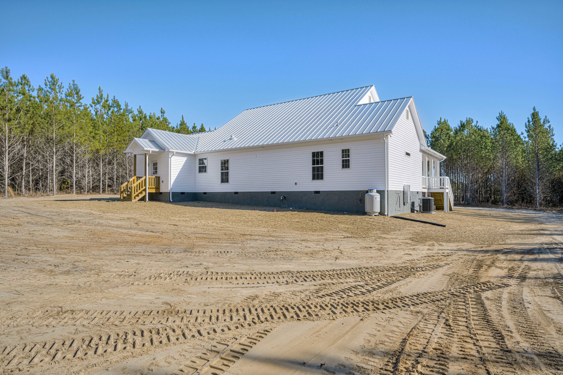 White house with metal roof, wooden exterior stairs, sandy dirt yard with tire tracks, large white container with blue label, trees in background, clear sky above