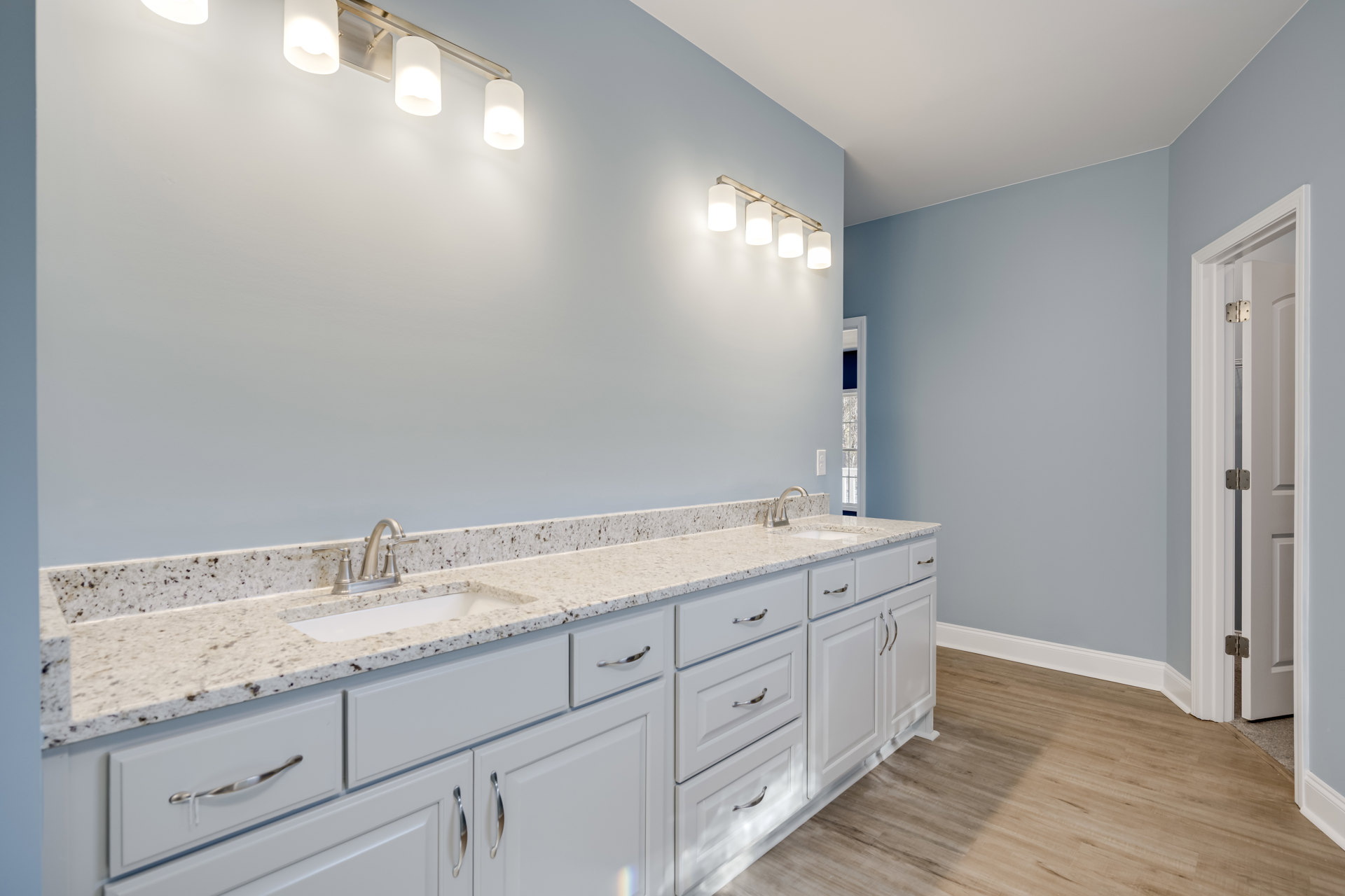White bathroom cabinets with marble countertops, wood flooring with white trim, wall-mounted light fixture with four bulbs, sink and chrome faucet, tile backsplash, drawers and