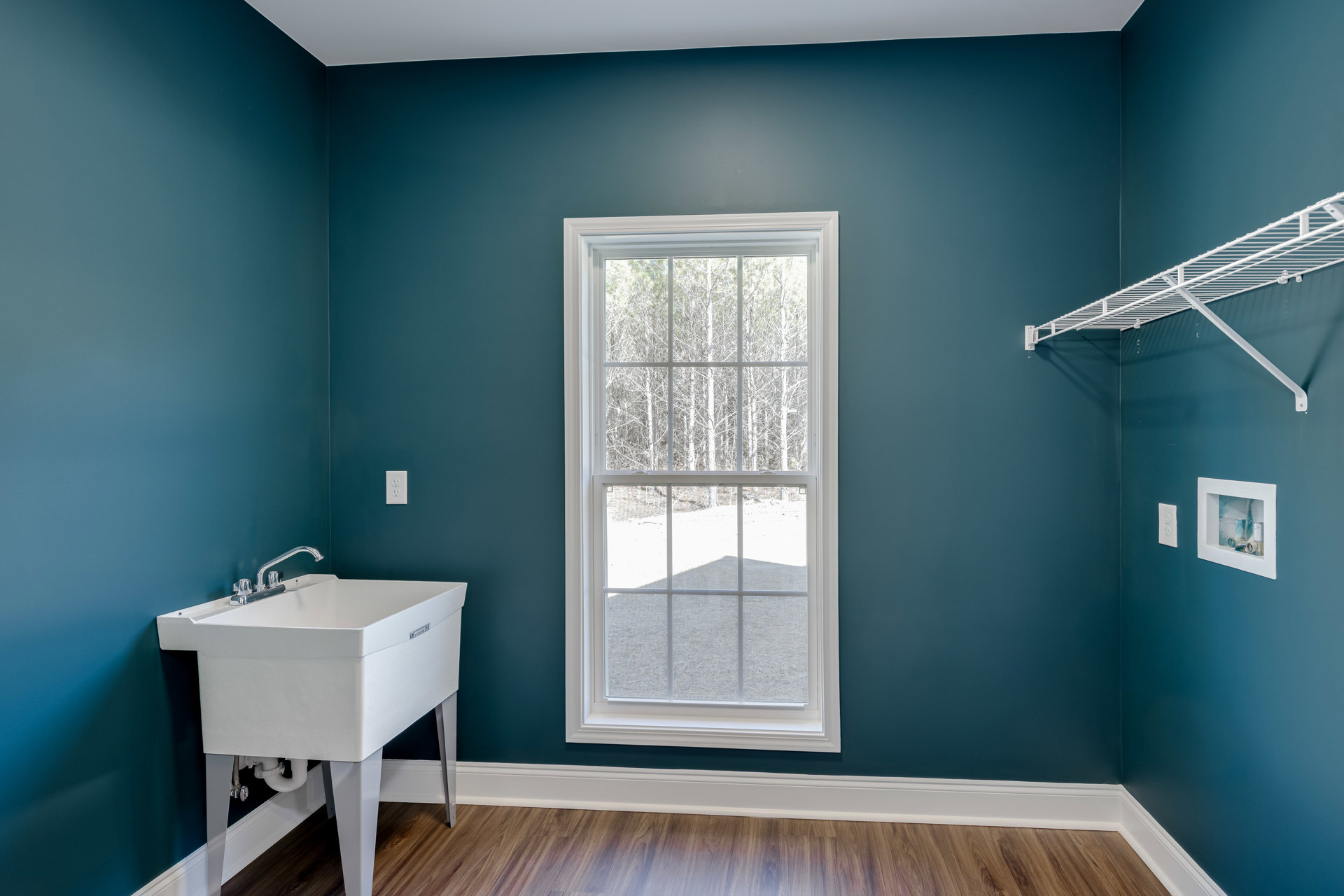 Bathroom with white pedestal sink, wood floor, window overlooking trees, white shelf mounted on wall, white ceiling.