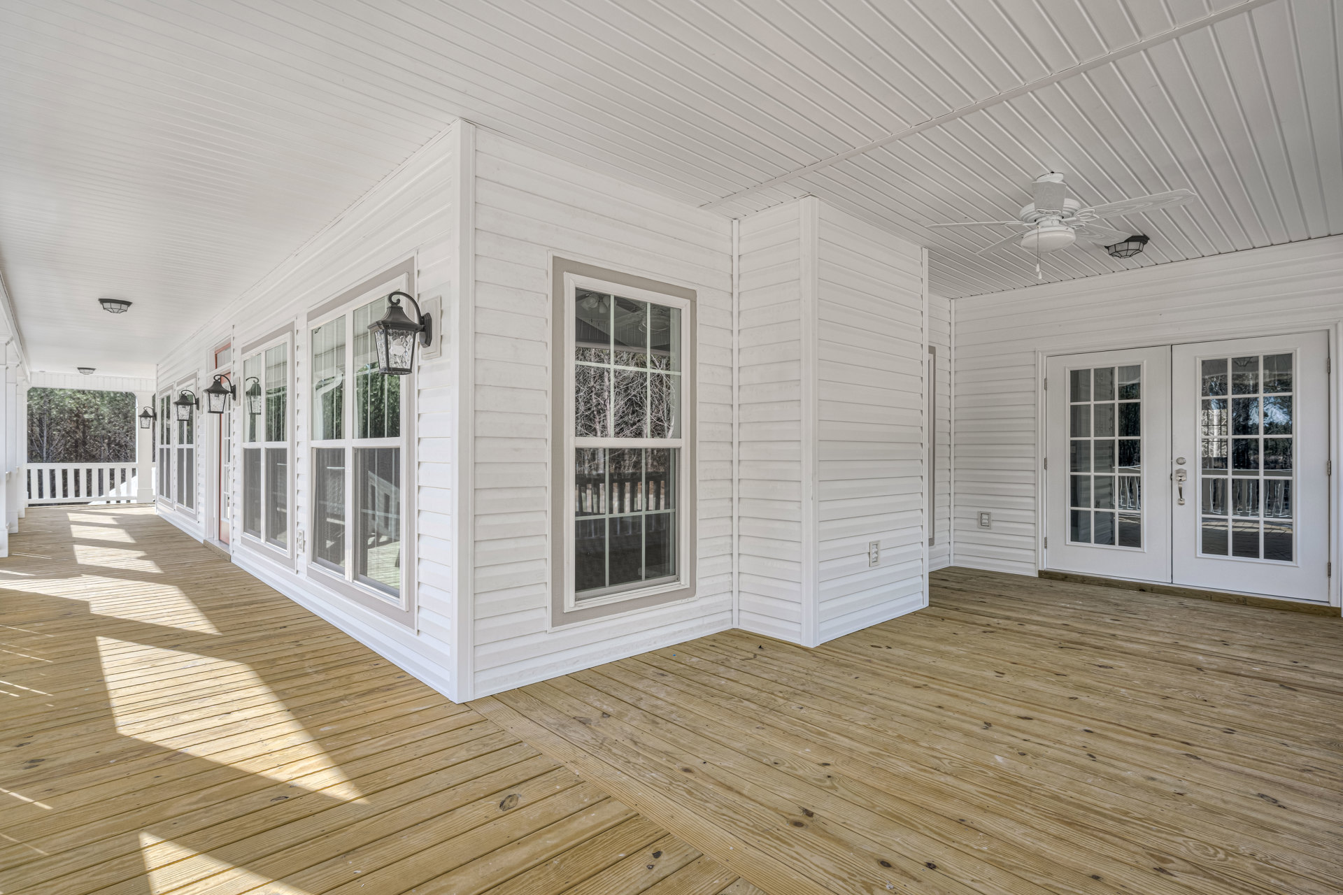 Open-concept interior with white walls, wood flooring, glass-paneled double doors, large window overlooking trees, wooden deck, and modern lamp fixture