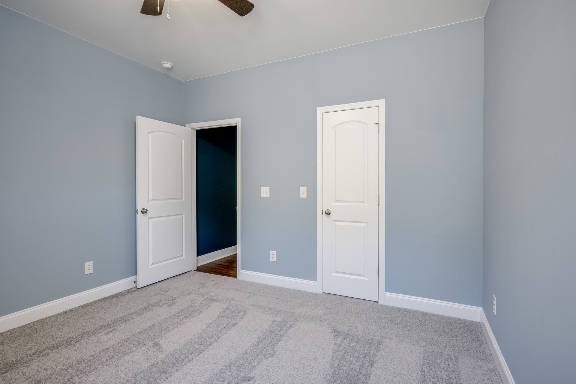 Carpeted room featuring two white doors with silver knobs, a ceiling fan with light, neutral walls, and a black framed board on one wall.
