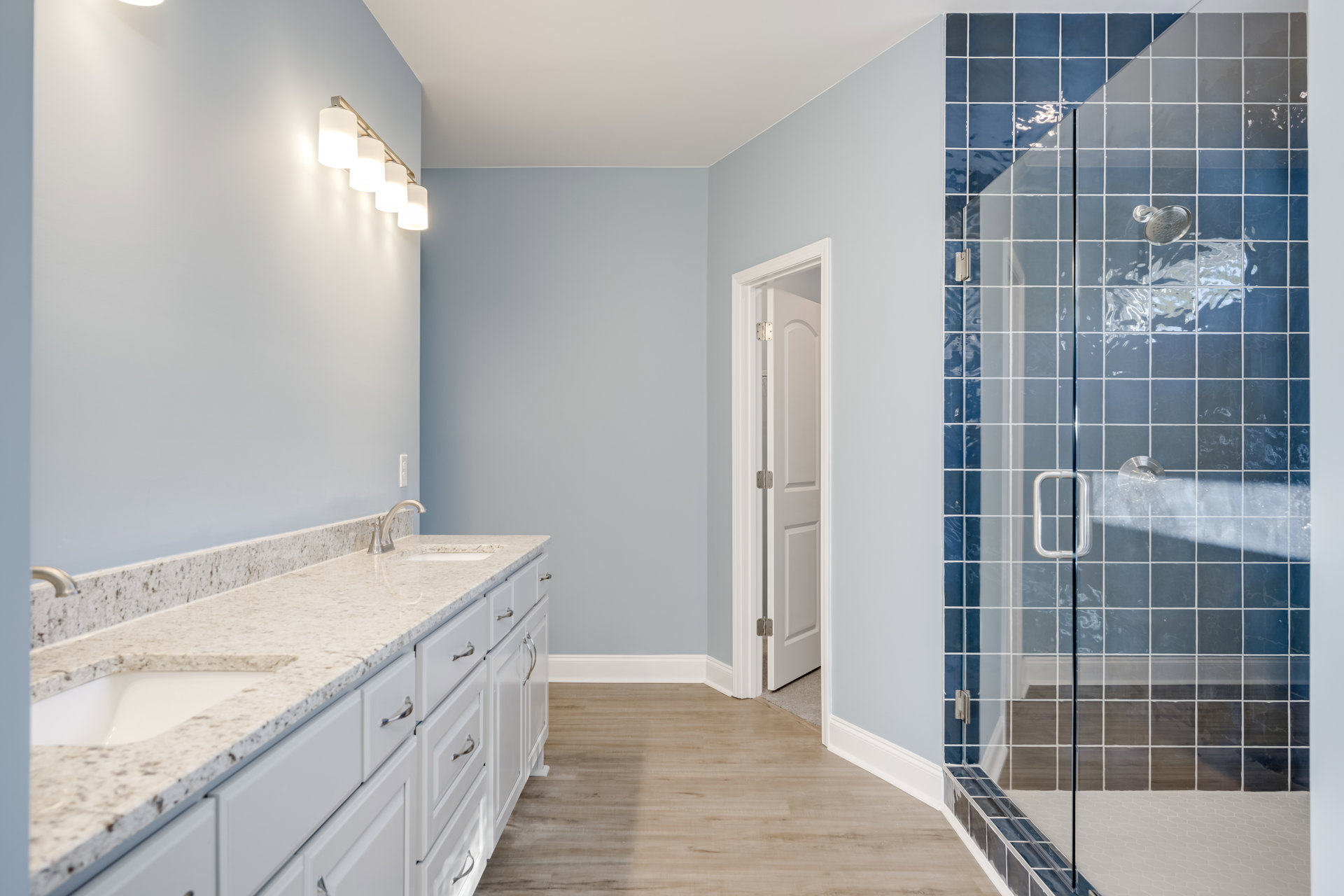 Bathroom with glass shower enclosure, white double sink vanity, wood flooring, white trim, and wall-mounted row of lights