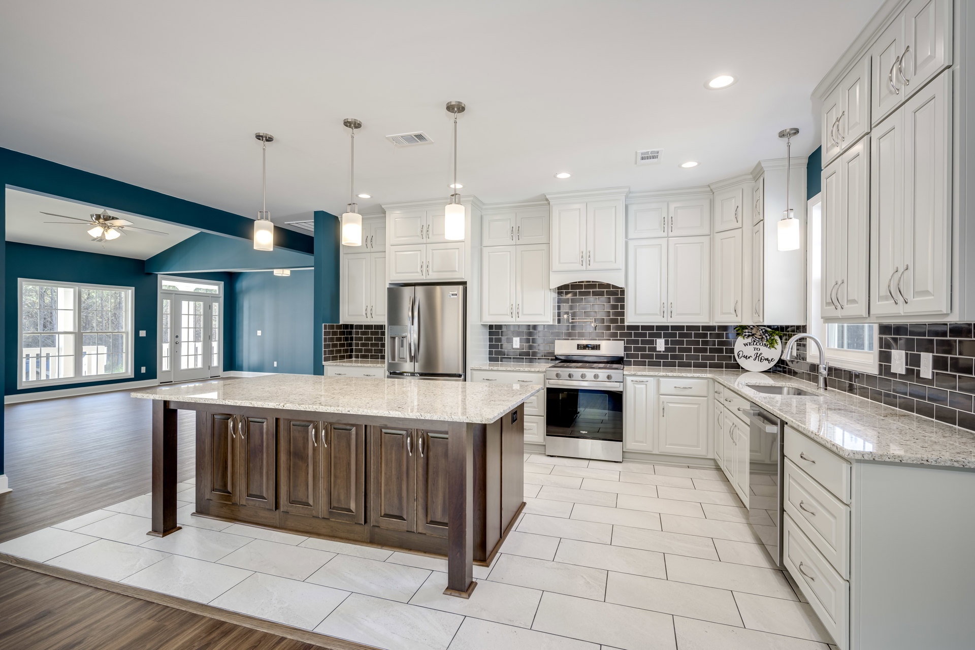 White kitchen cabinets, granite island with marble veining, silver refrigerator, black tile countertop, window with white trim, white door with glass panes, decorative sign with