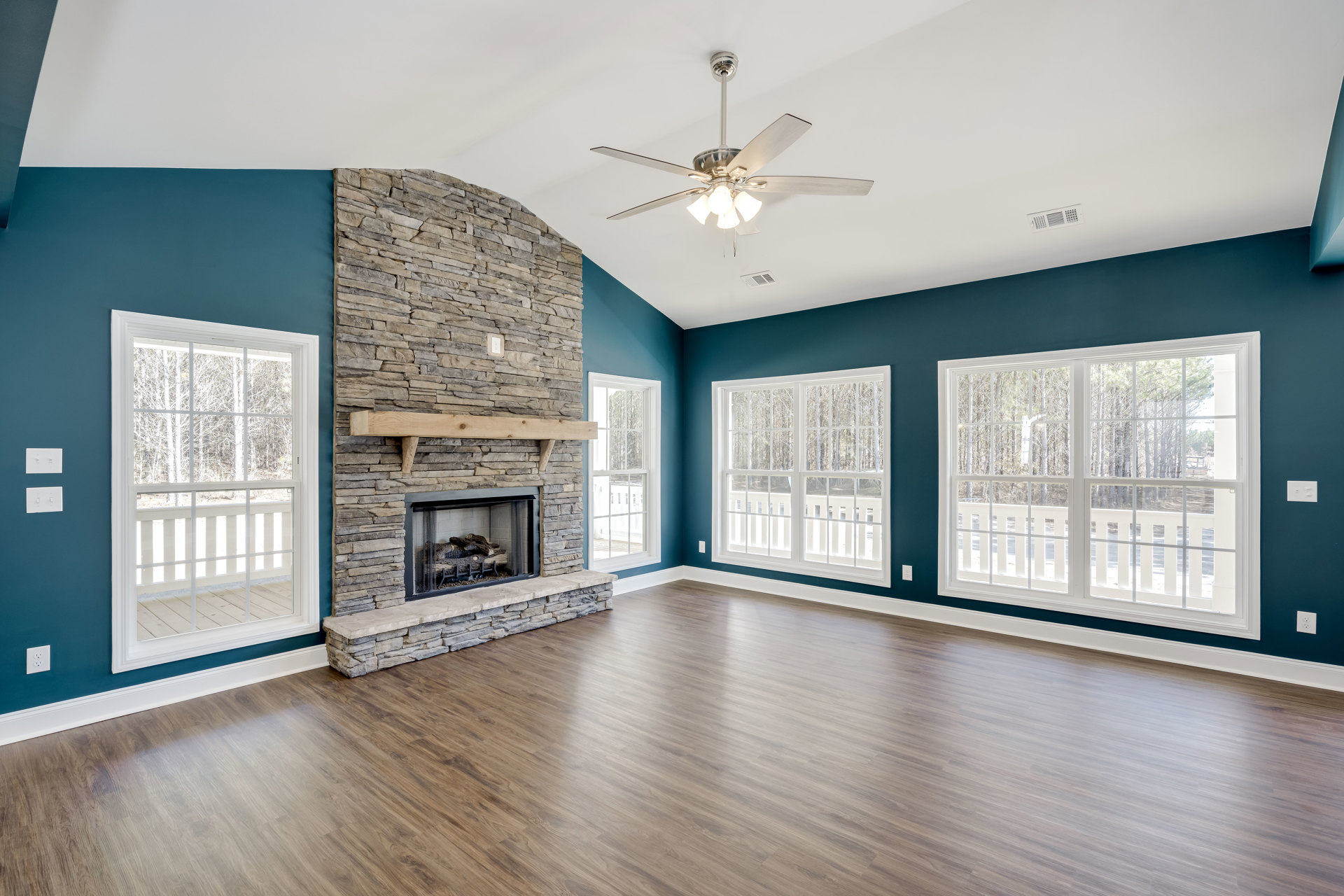 Living room with wood flooring, white fireplace featuring a wood mantel and mesh screen, ceiling fan with light fixture, large window overlooking trees.