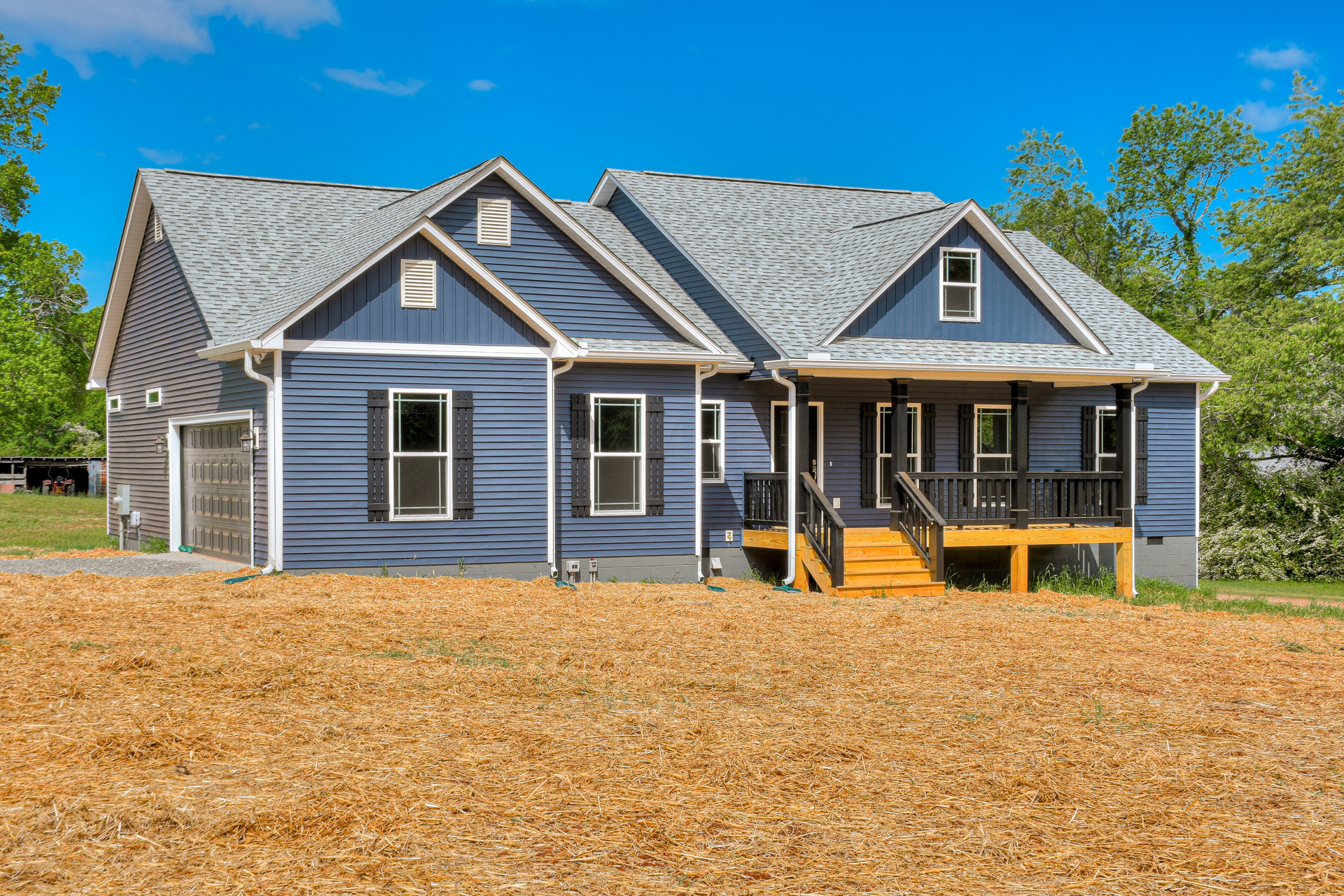 Blue siding house with white trim, wooden porch and stairs, straw-covered yard, mature trees in background