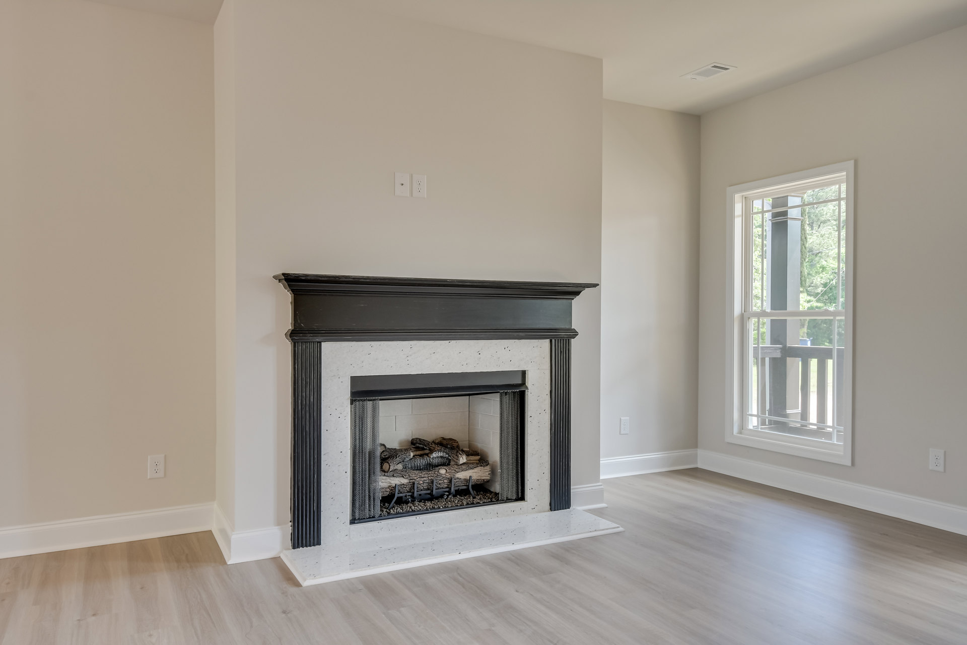 Wood burning fireplace with black frame, stacked logs and rocks inside, light-colored flooring, large window with railing and view of trees, white walls with crown molding
