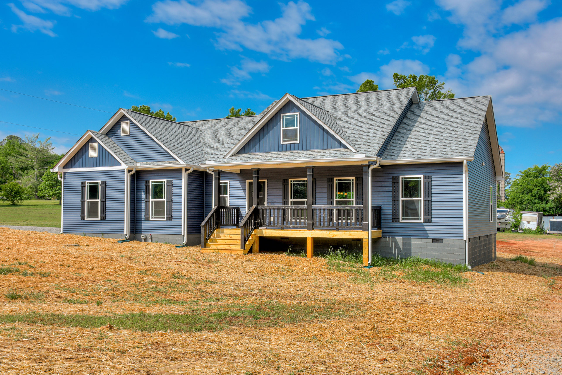Two-story house with white siding, covered front porch with stairs, large grassy yard, white-framed windows, gabled roof, and mature trees in the background