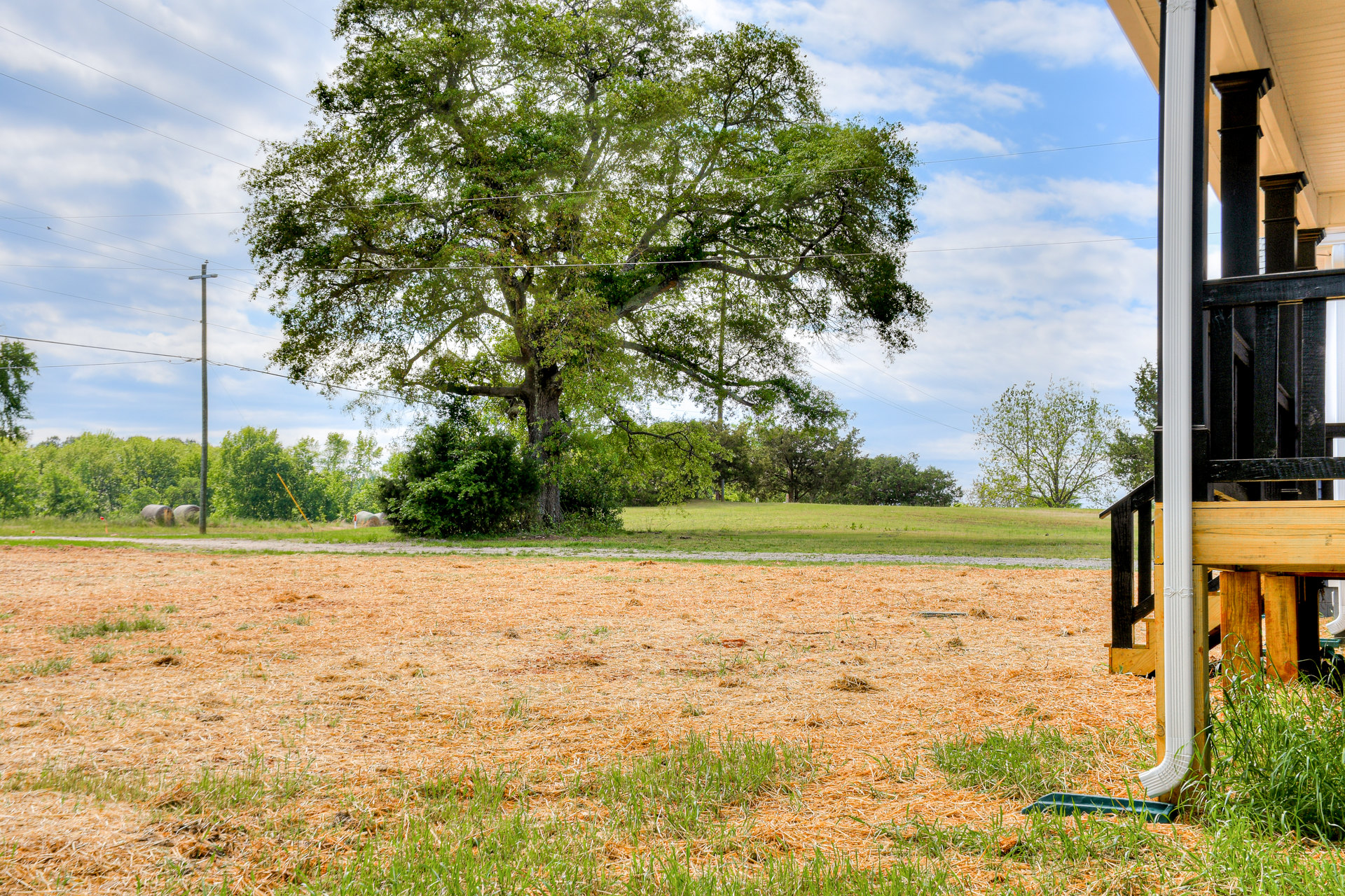 Modern deck with black metal railing overlooking grassy field, large tree, wood fence, telephone pole, and two hay bales under partly cloudy sky